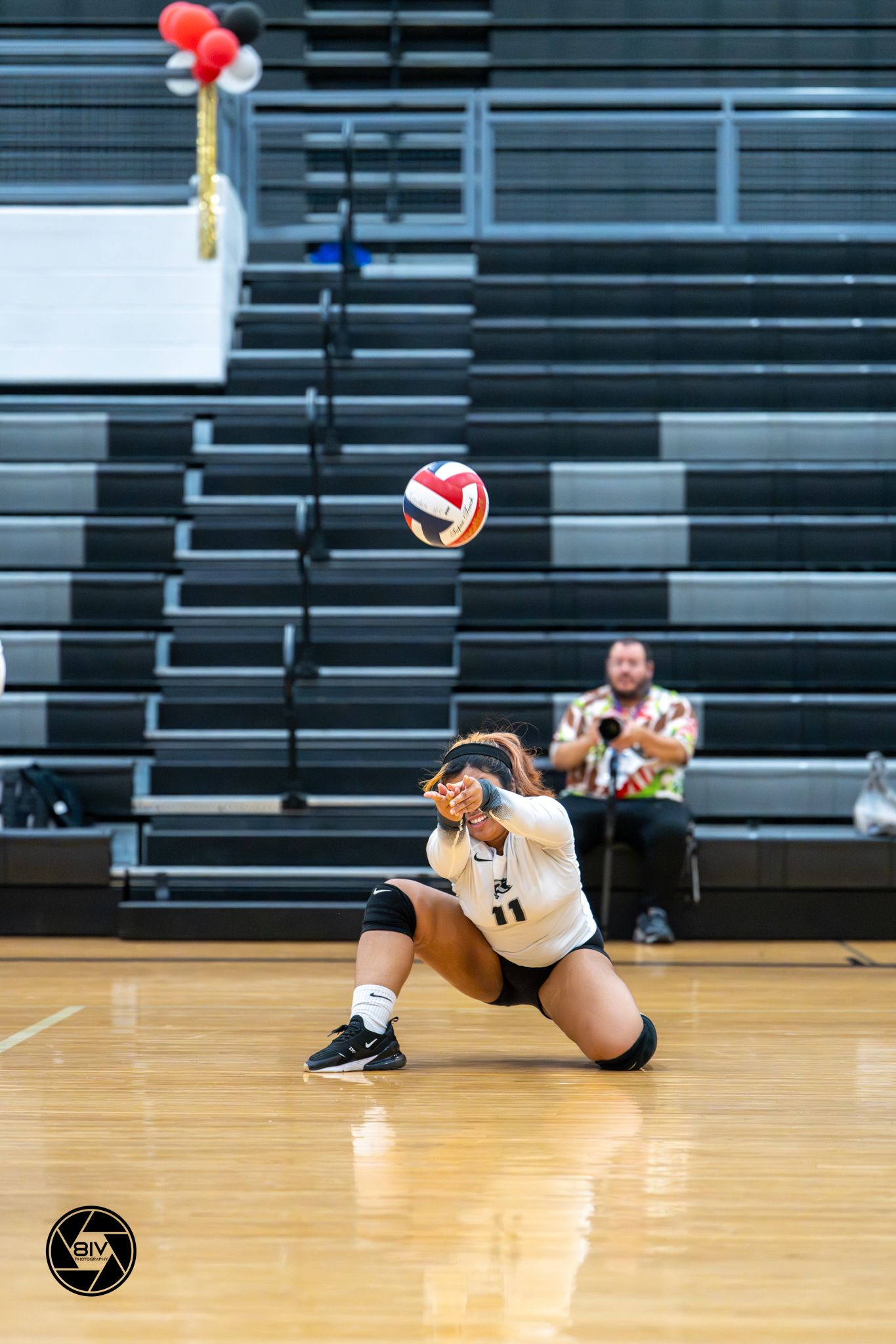 Female volleyball player performs a low dig on indoor court, wearing black and white uniform with ball in mid-air.