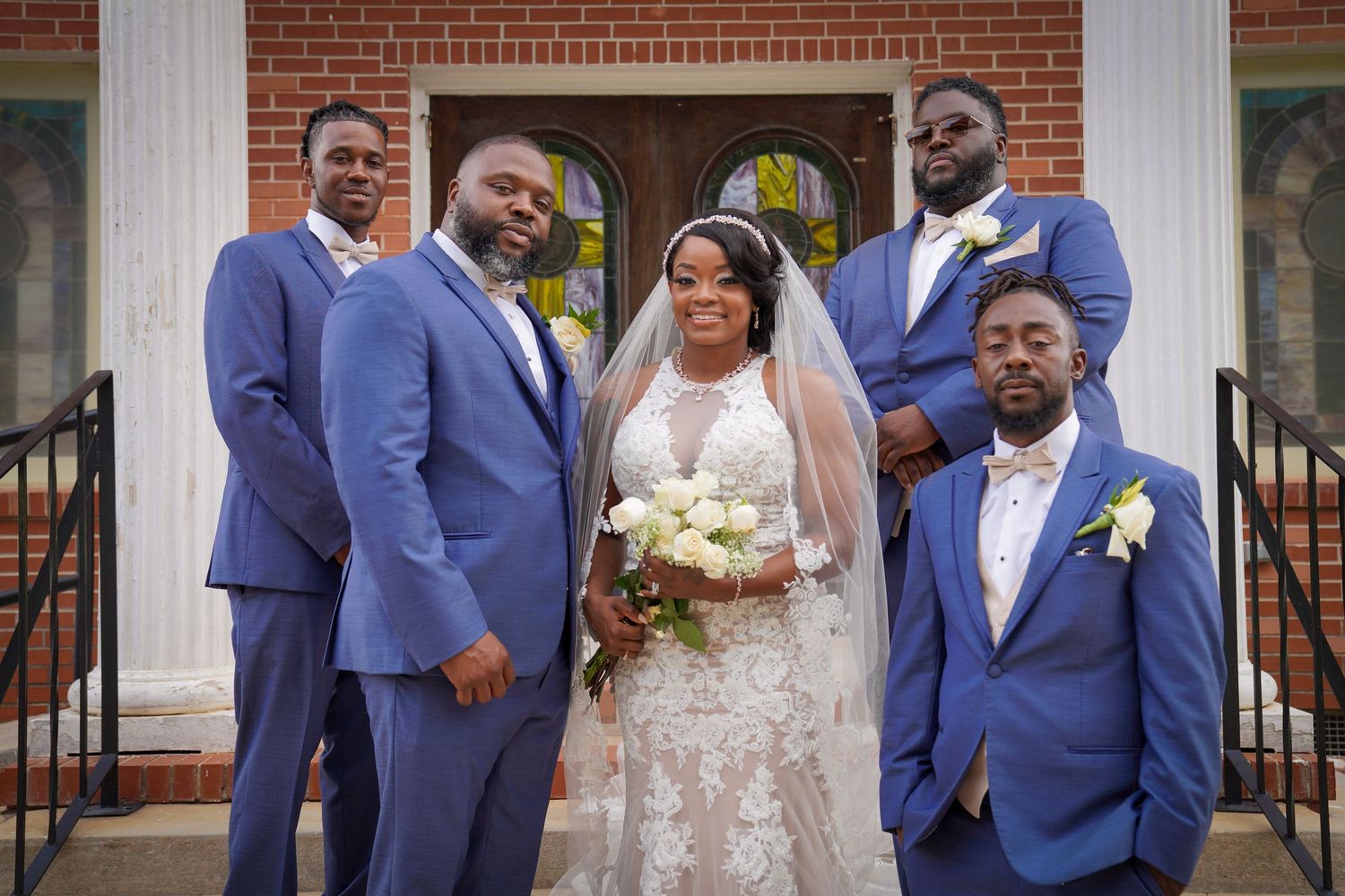 Bride in white lace gown with veil poses with four groomsmen in blue suits outside a brick church.