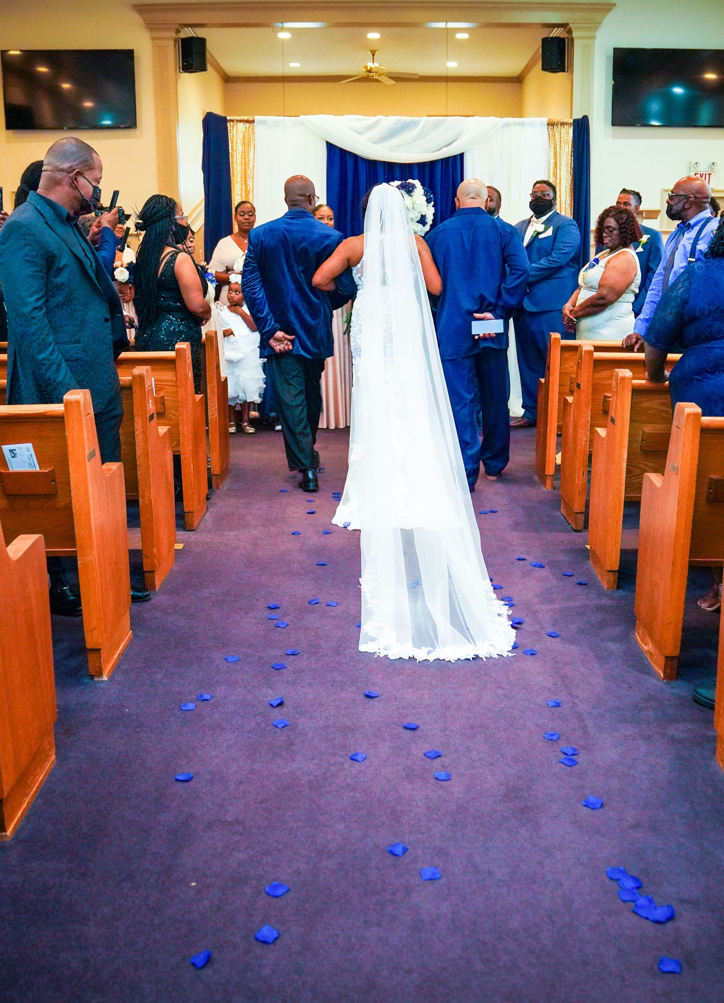 Bride walking down church aisle with blue rose petals, long white veil trailing, guests in navy attire lining wooden pews.