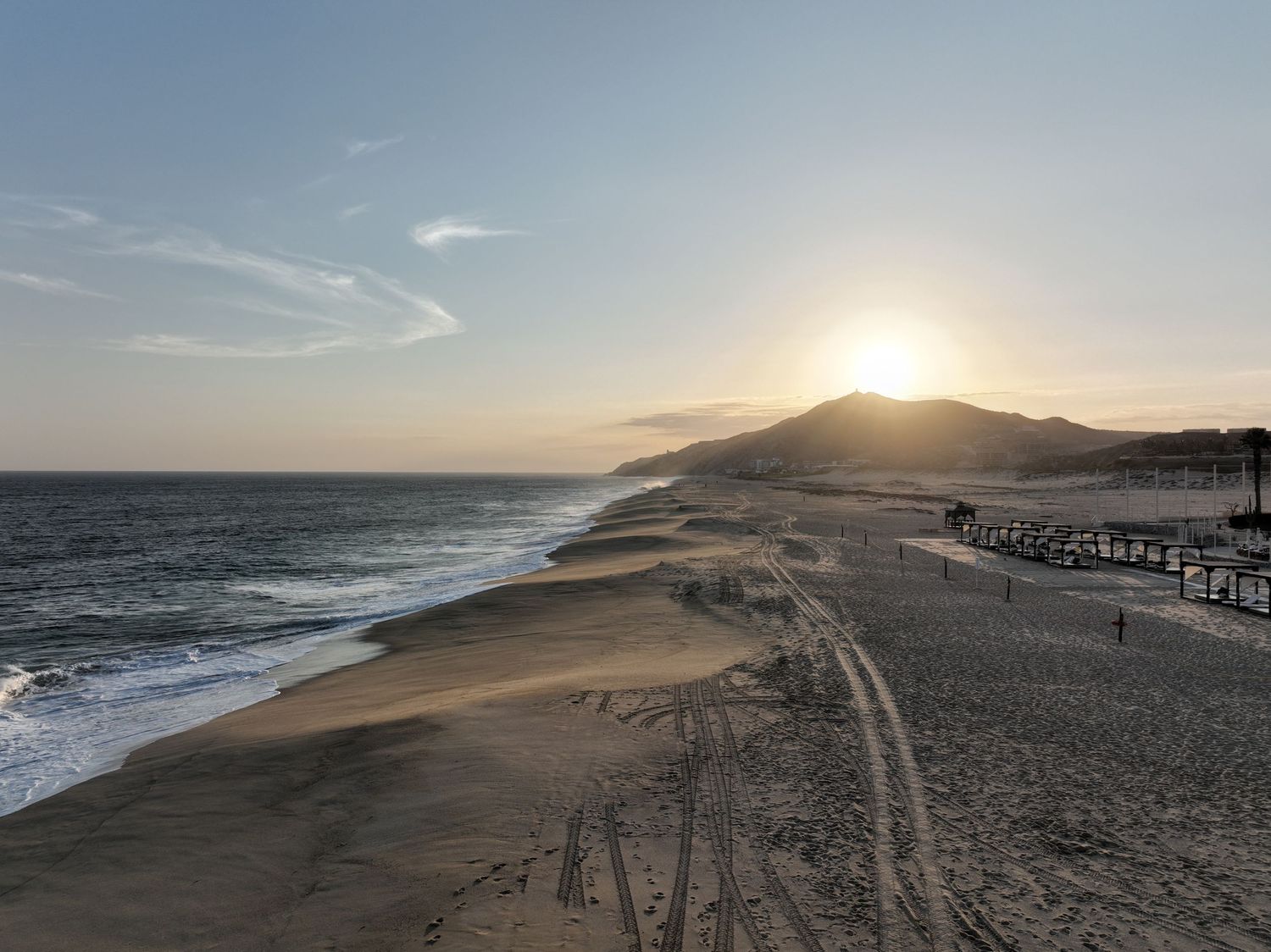 Aerial view of a sandy beach at sunset with golden light over a coastal hill and calm ocean waves.