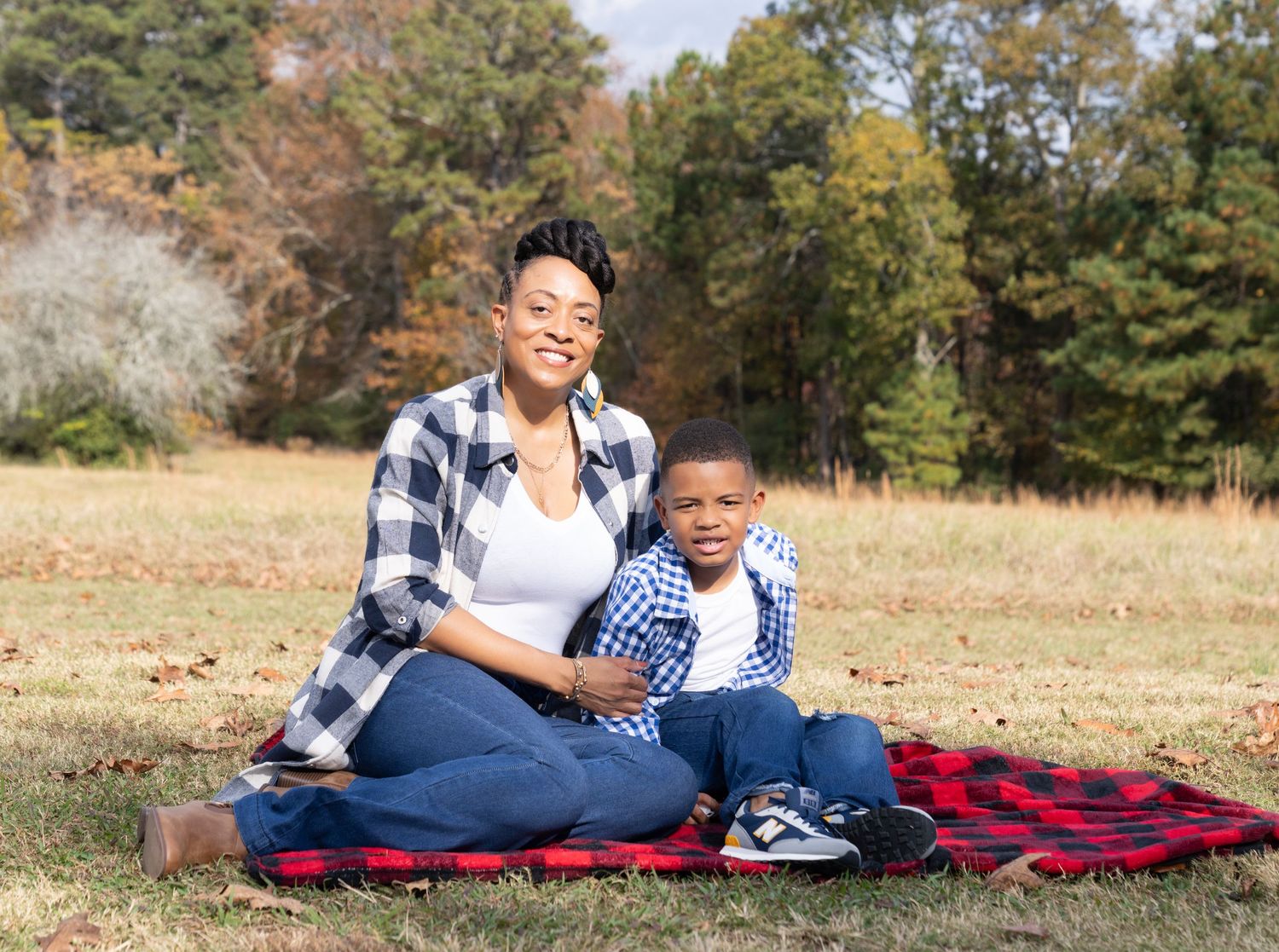 A woman and young boy smile while sitting on a red plaid blanket in a grassy park with autumn trees behind them.