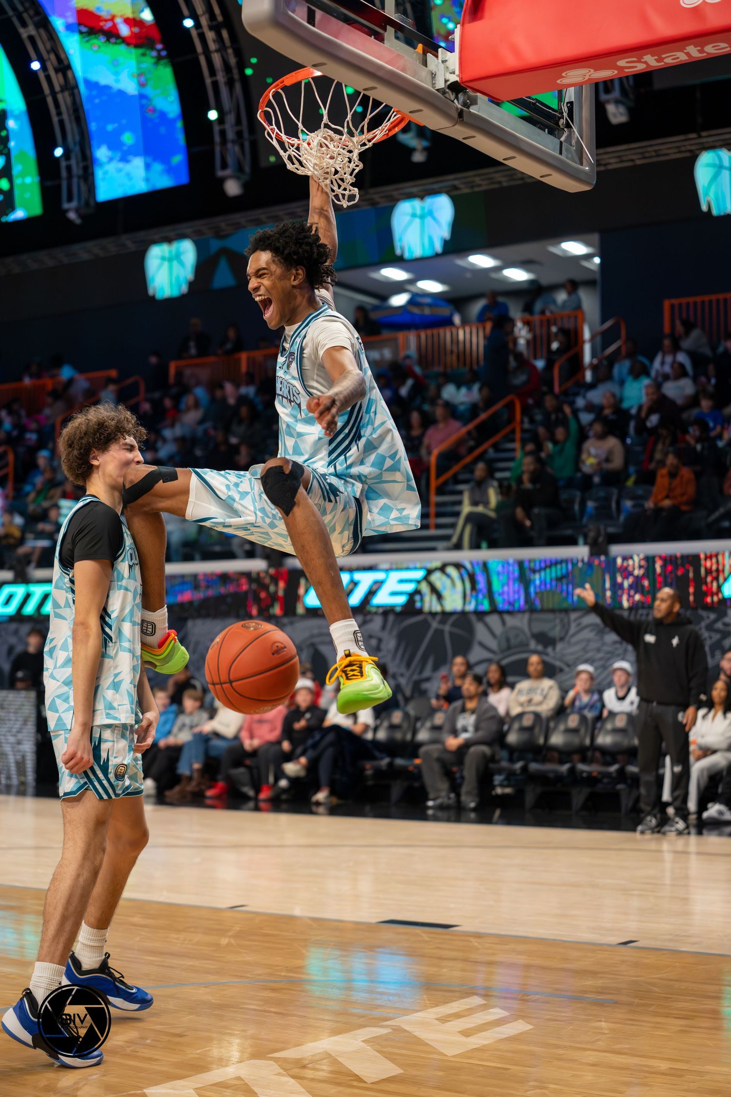 Basketball player hangs on rim after dunk during Overtime Elite game as teammate watches below.