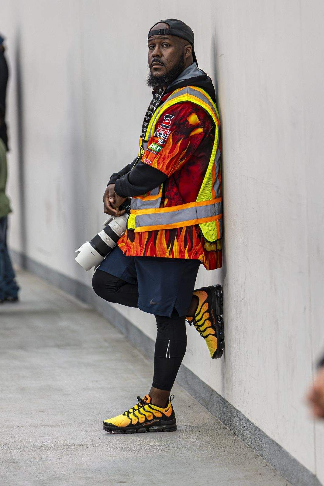 Man in flame jacket, safety vest, yellow Nike sneakers leans against wall holding camera lens.