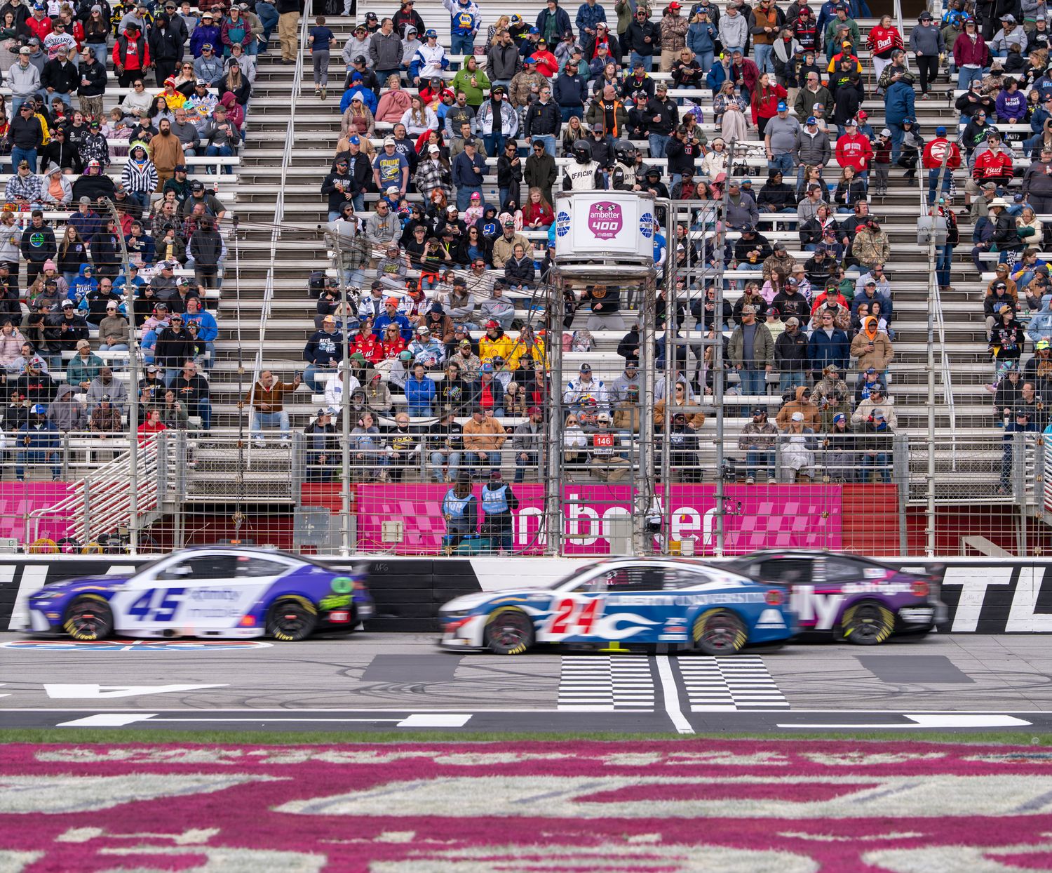 NASCAR race cars speed past the finish line at a packed Echo Park Speedway oval track with cheering fans filling the grandstands.