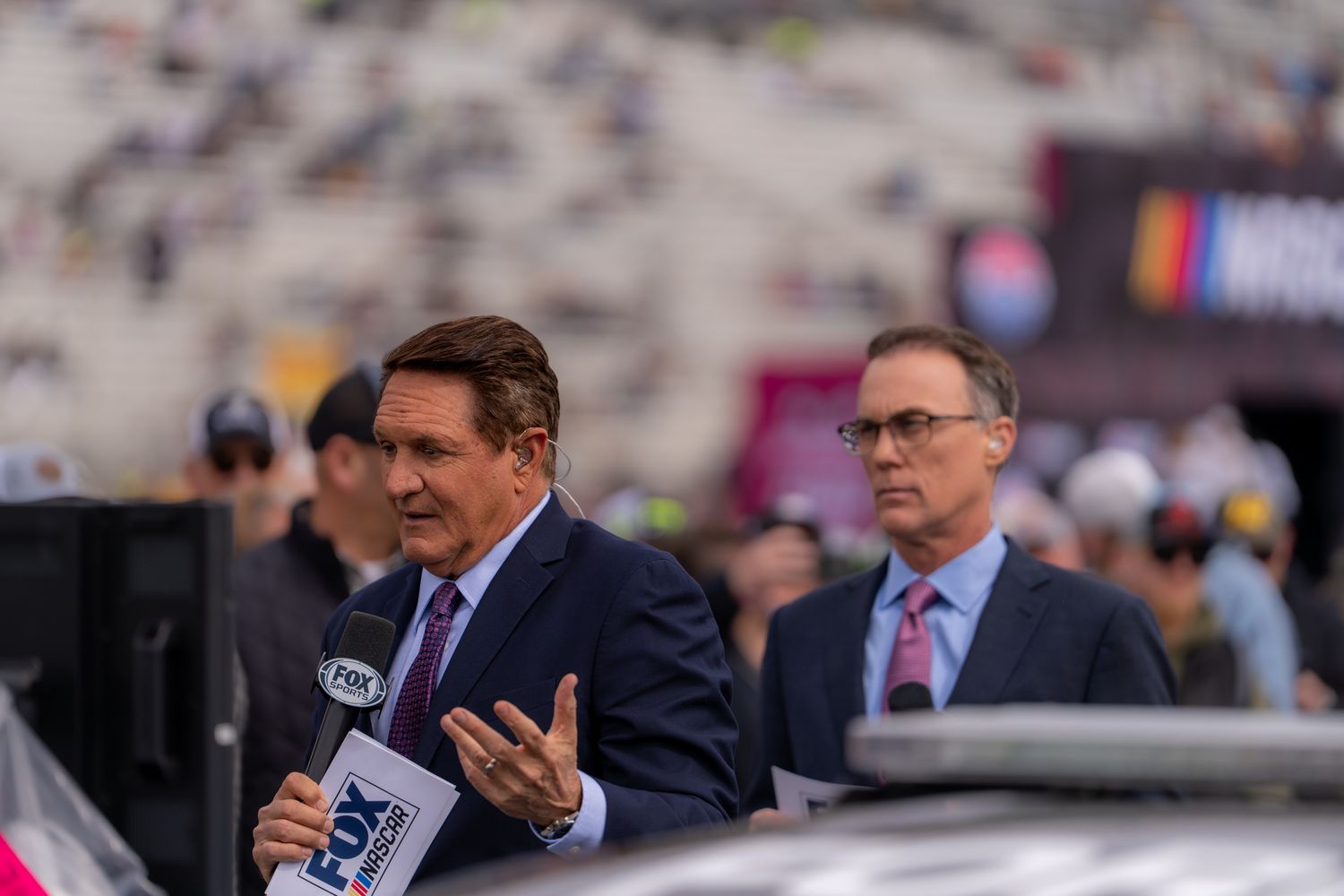 Two FOX Sports broadcasters speaking at a NASCAR event with grandstands visible in the background.