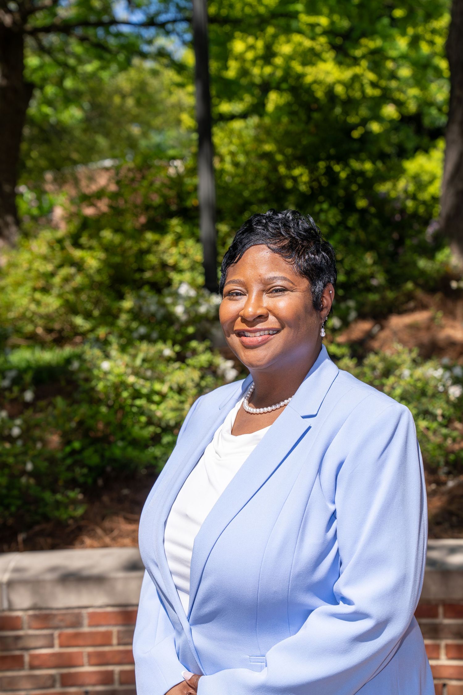 Professional woman in light blue blazer and pearl necklace smiling outdoors near brick wall and green trees.