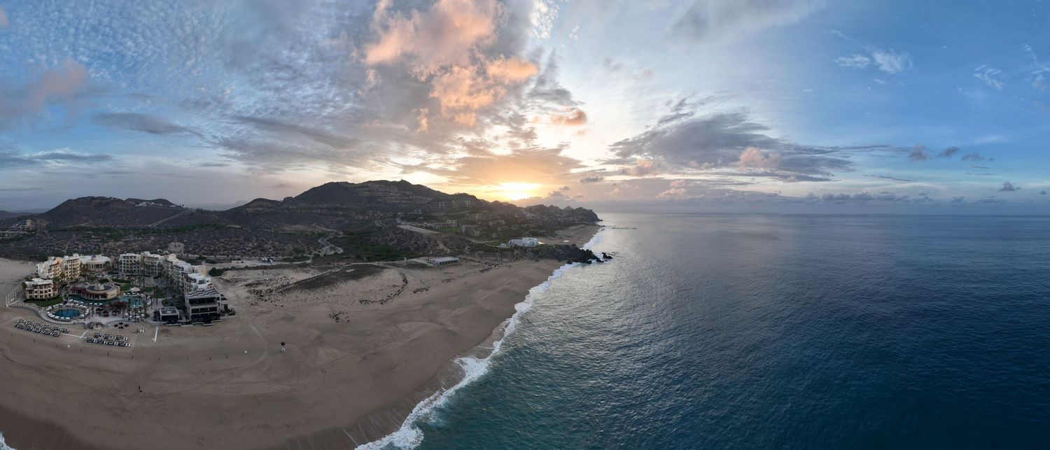 Aerial panoramic view of a sandy beach coastline at sunset with mountains, resort, and calm blue ocean waters.