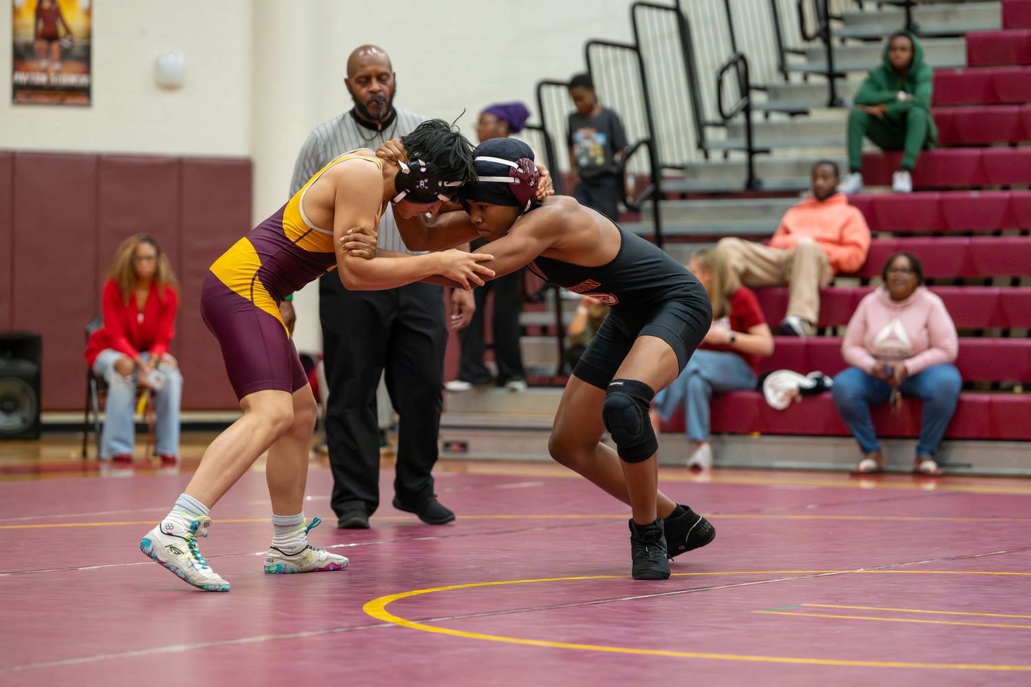 Two wrestlers compete on a mat during a high school wrestling match in a gymnasium with spectators watching.