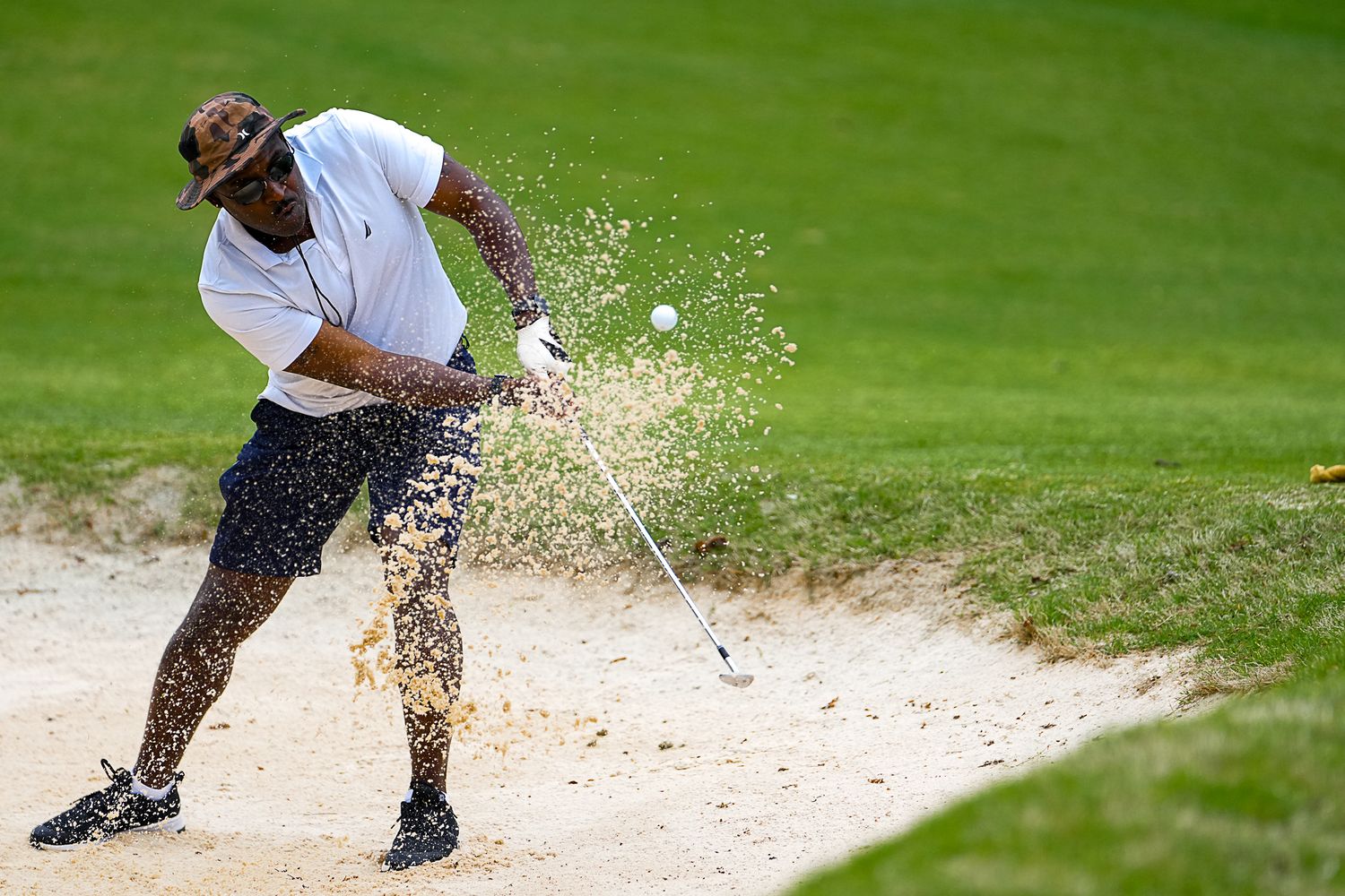 Golfer hitting a bunker shot from a sand trap, sending sand flying on a golf course.