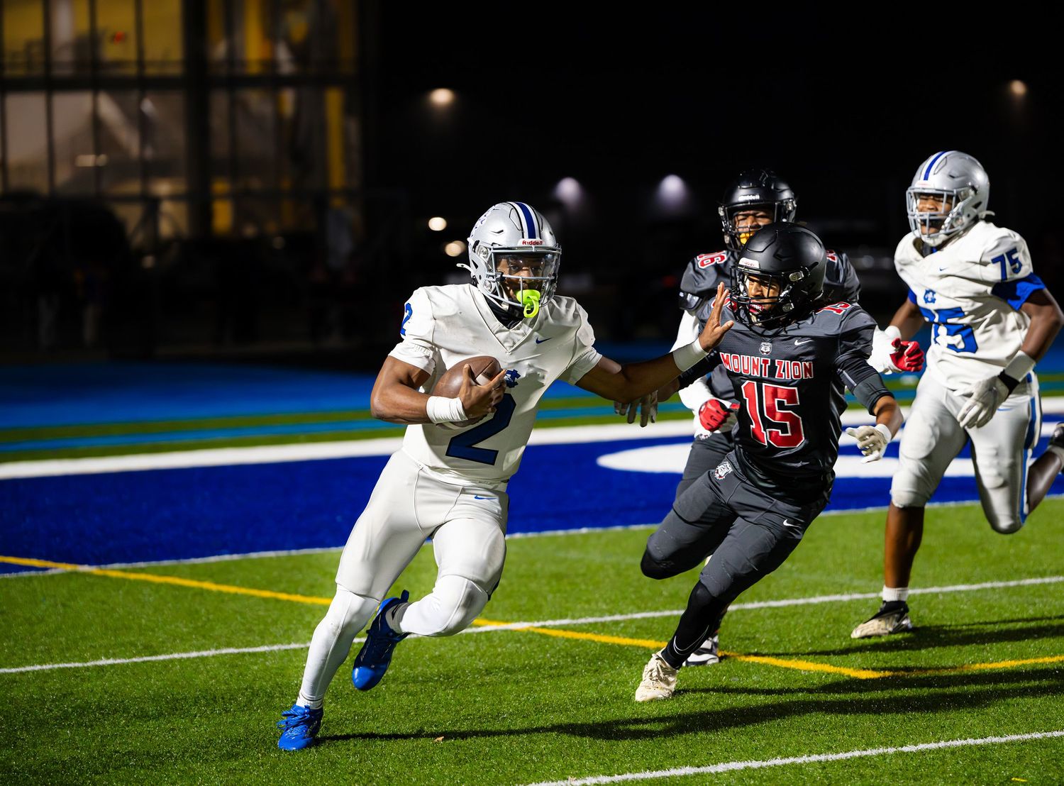 High school football, Justin Murphy  in white uniform runs with ball while defender in red jersey number 15 attempts tackle at night game.