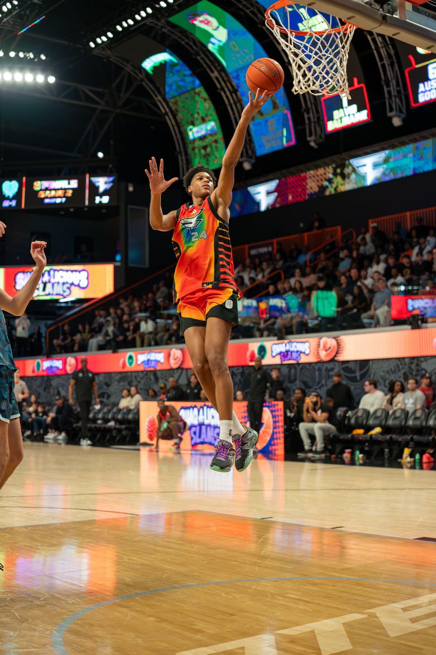 Basketball player in orange uniform leaps toward hoop during colorful indoor game with crowd watching.