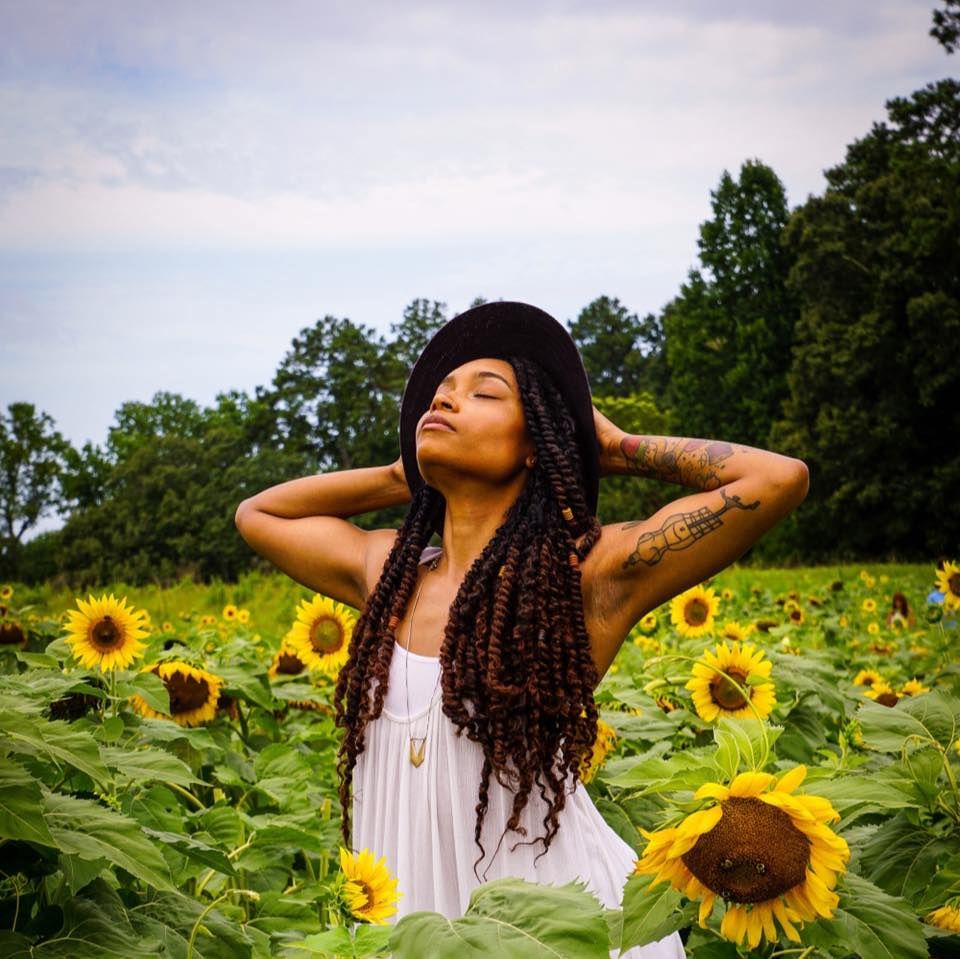 Woman with braids and black hat poses in a vibrant sunflower field, wearing a white dress with arm tattoo.