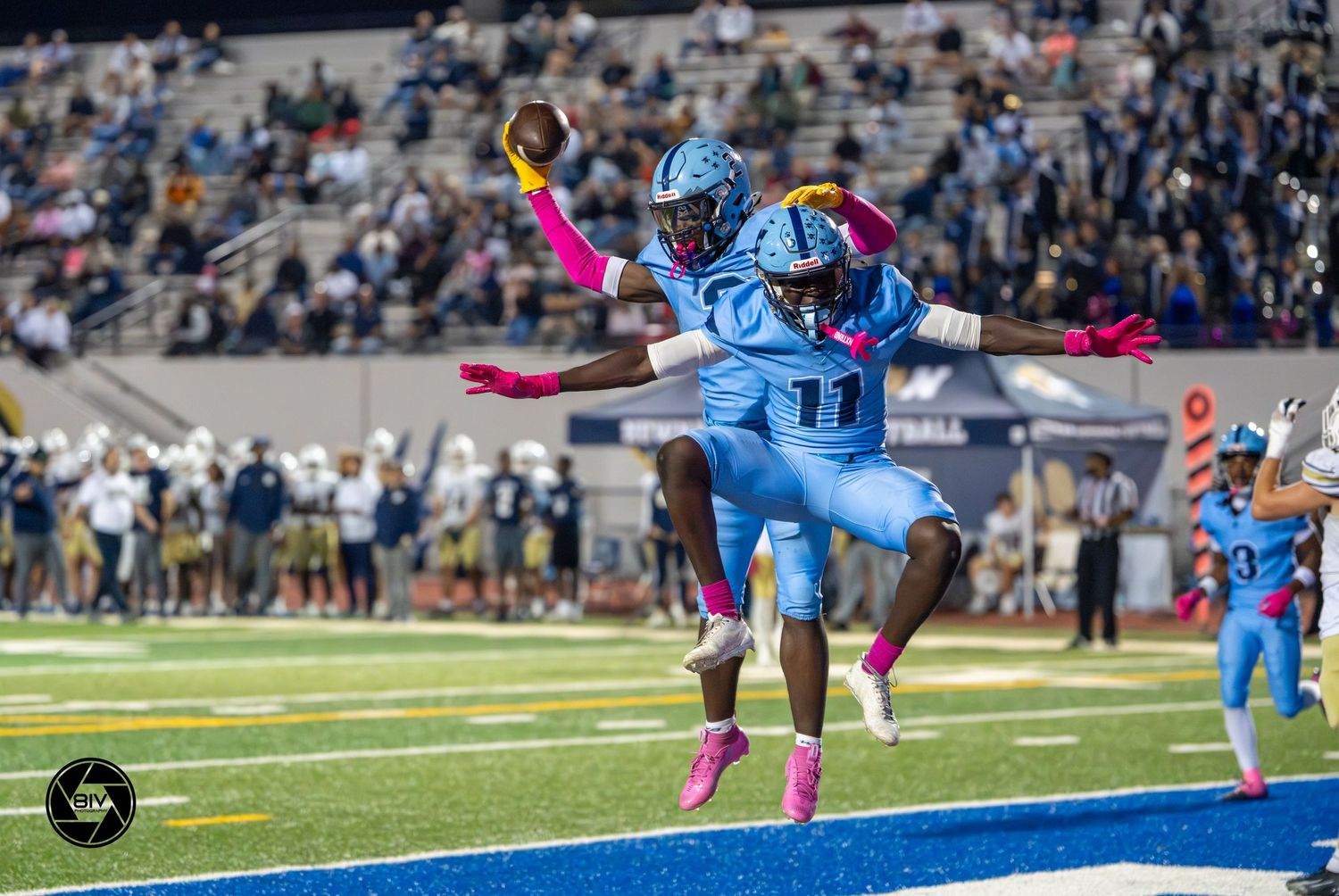 Two football players in blue uniforms celebrate a touchdown in the end zone during a night game.
