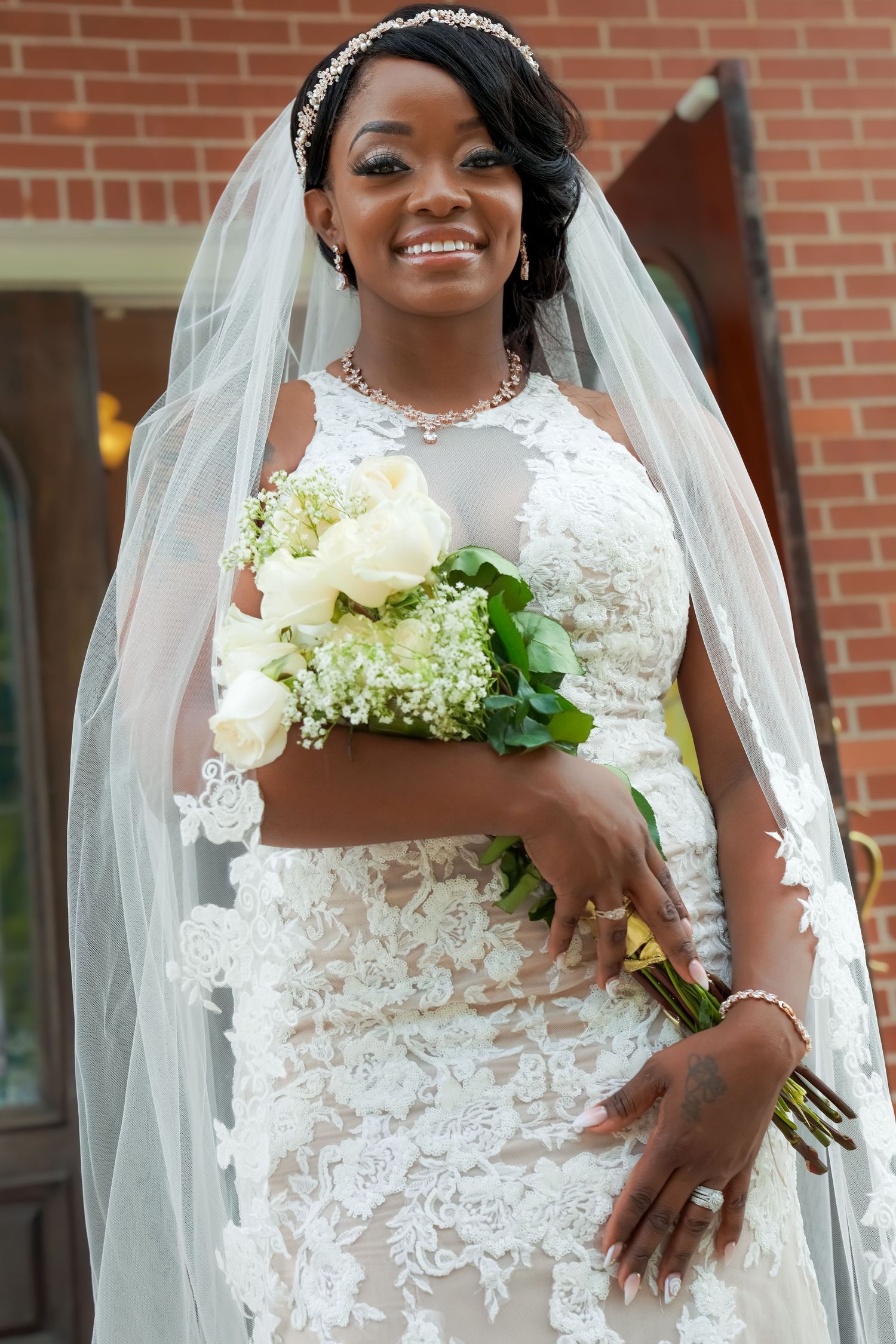 Smiling bride in lace wedding dress and veil holds white rose bouquet outside brick church.