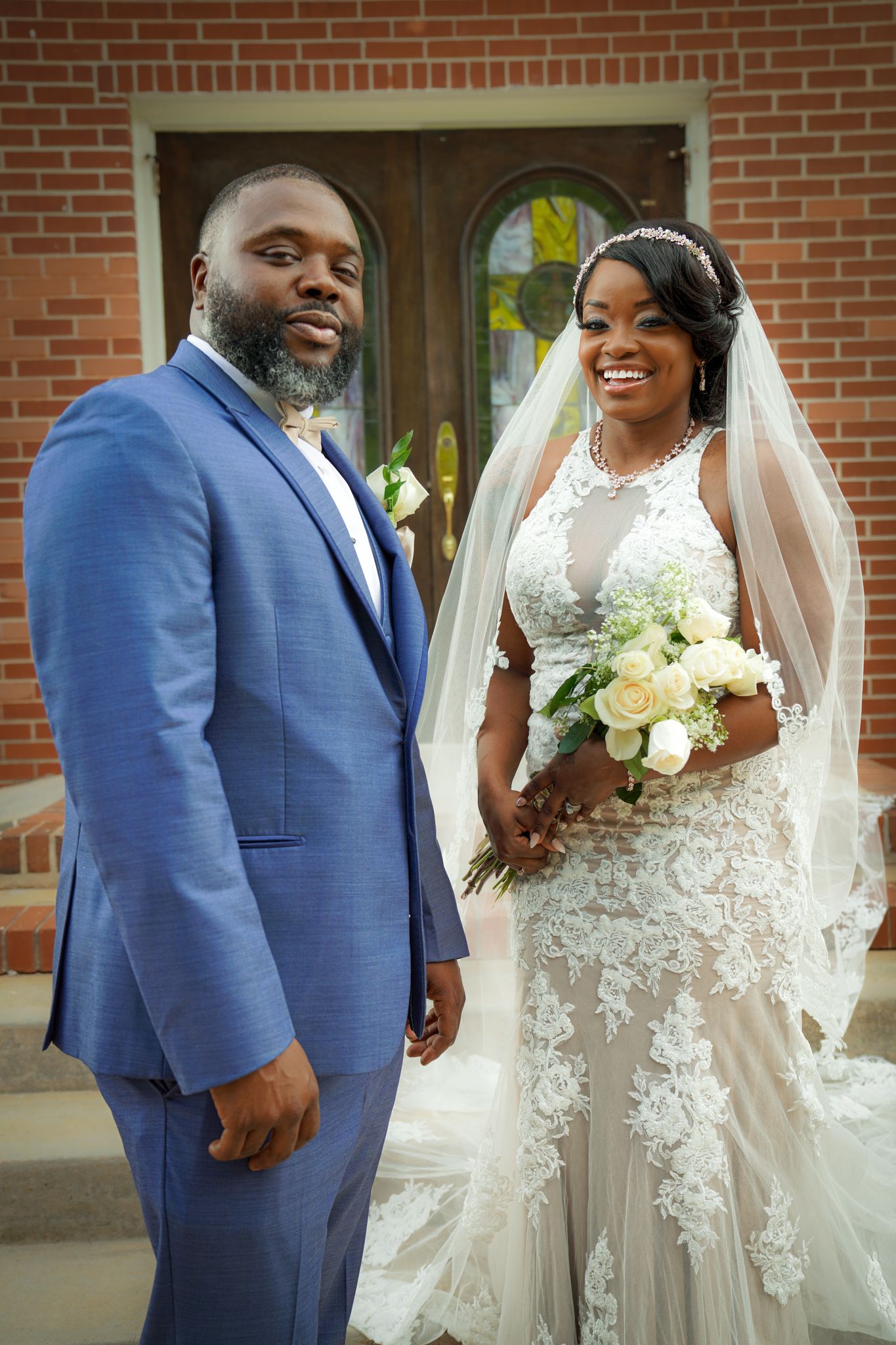 A smiling bride in lace gown with veil holds white bouquet beside groom in blue suit outside brick church.