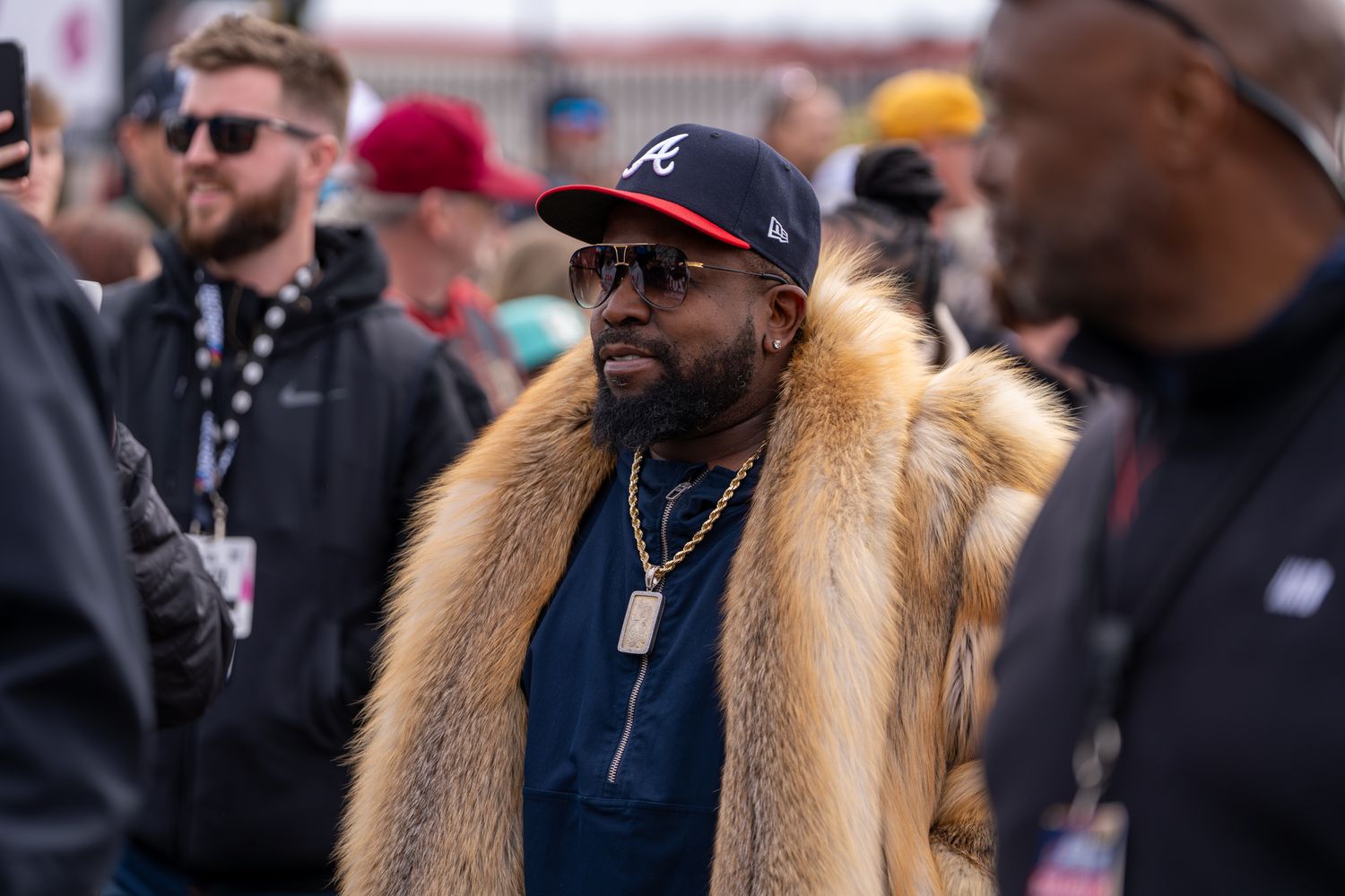 Big Boi from Outkast in fur coat and Atlanta Braves cap with gold chain stands in crowd at outdoor event.