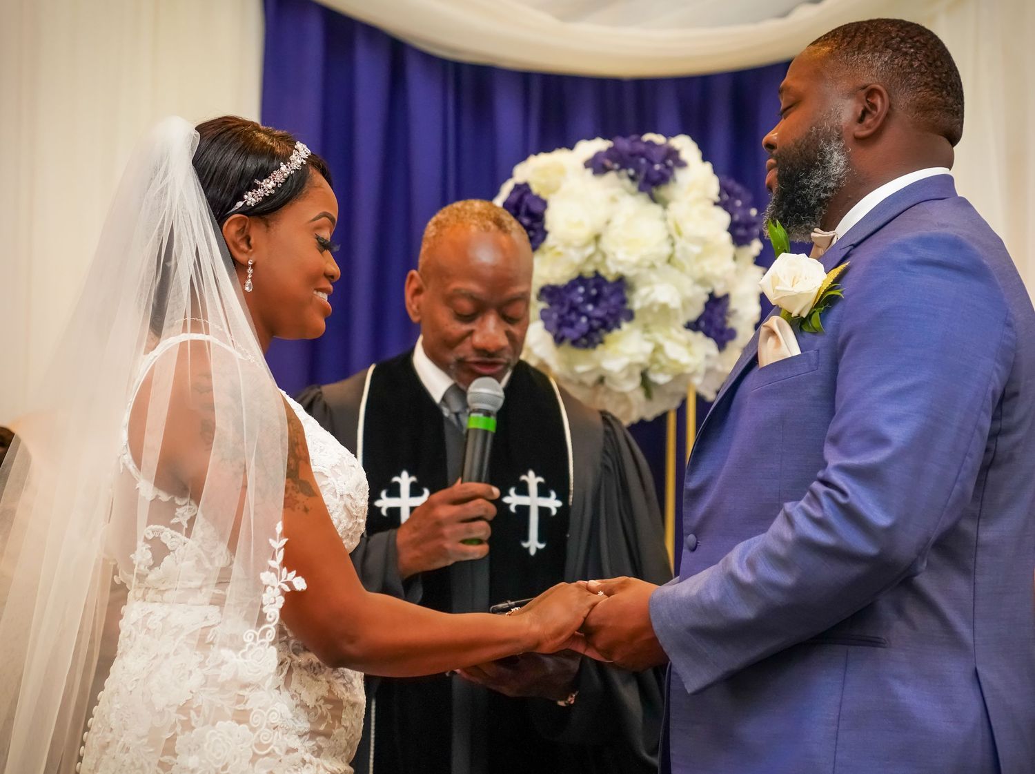 Bride and groom exchange vows at wedding ceremony with officiant holding microphone, purple and white floral backdrop.