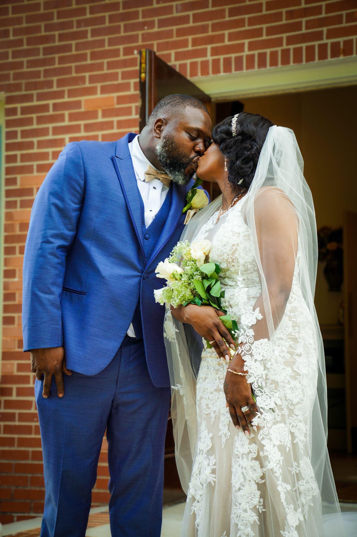 A bride and groom share a romantic kiss outside a brick church, she holds white flowers wearing a lace gown and veil.
