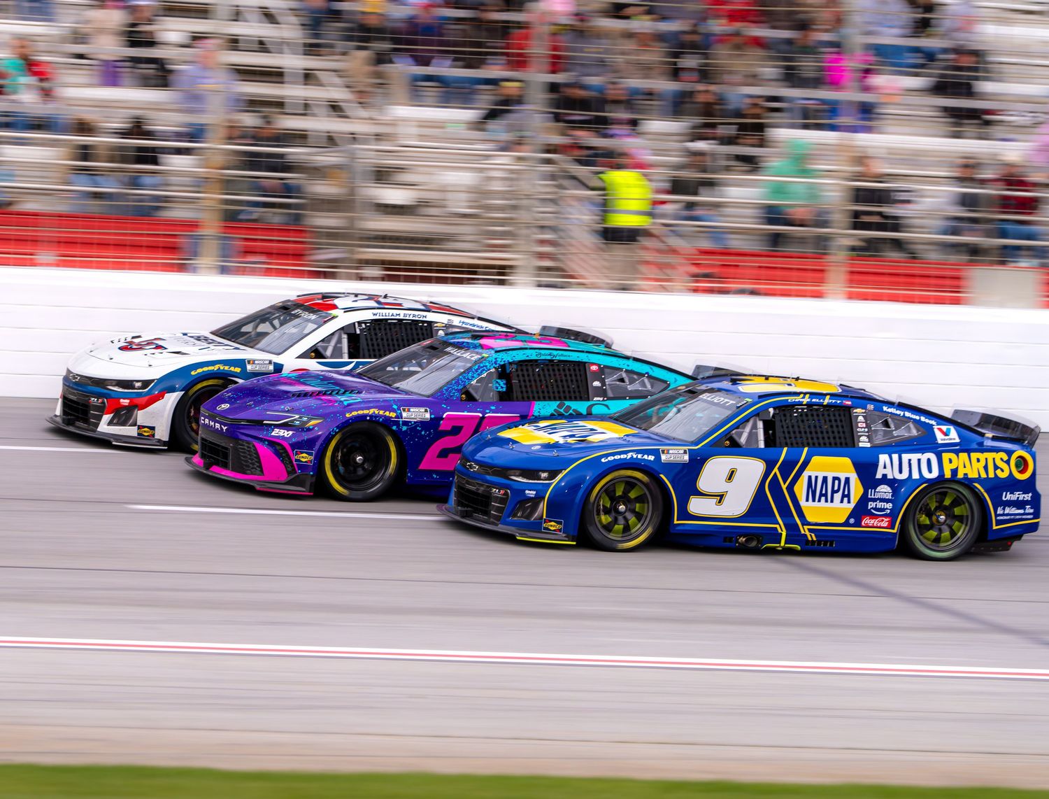 Three NASCAR race cars, including the NAPA Auto Parts No. 9, compete side by side on a banked oval track.