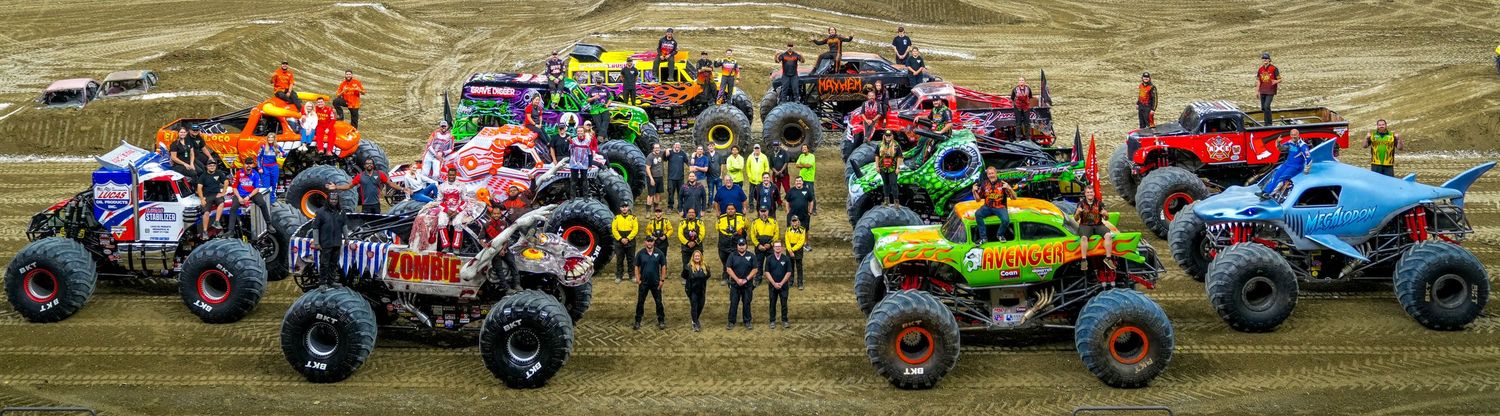 A lineup of colorful monster trucks with their drivers posing together on a dirt track at a Monster Jam event.