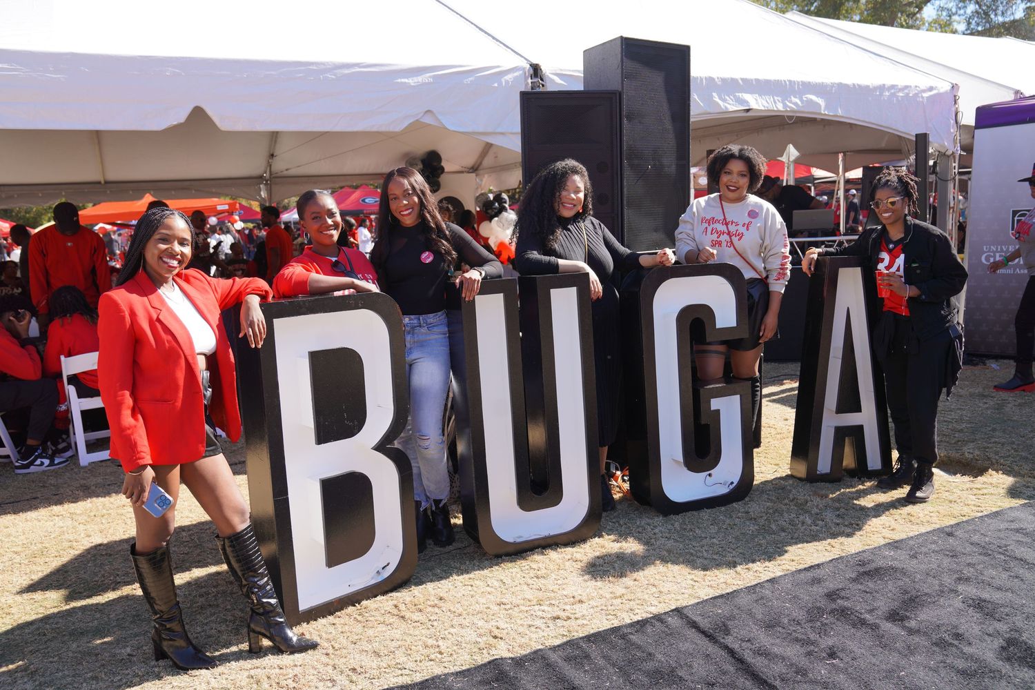 Group of women posing with large BUGA letters at an outdoor festival event, wearing red and black outfits.