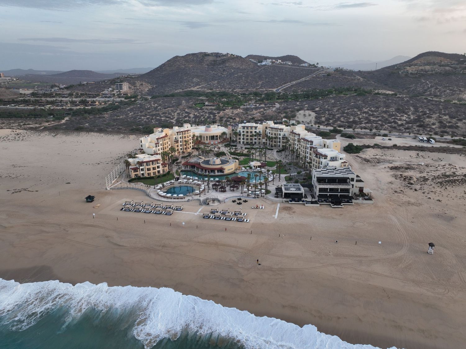 Aerial view of a beachfront resort complex on sandy shores with ocean waves, mountains, and desert landscape in Cabo San Lucas.