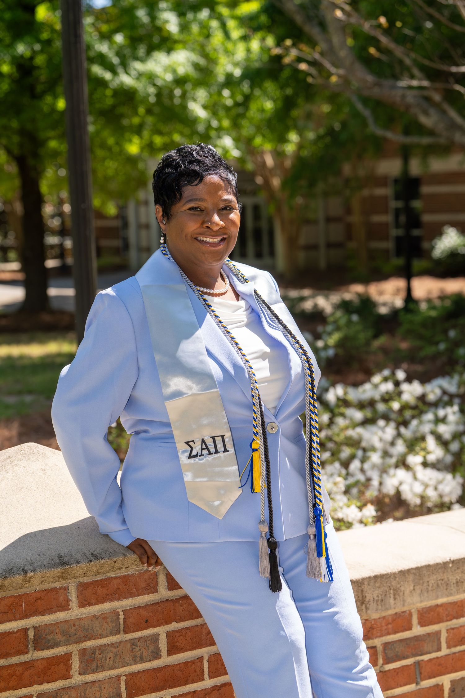 A smiling woman in a light blue suit wearing a Sigma Alpha Pi graduation stole and honor cords poses outdoors.