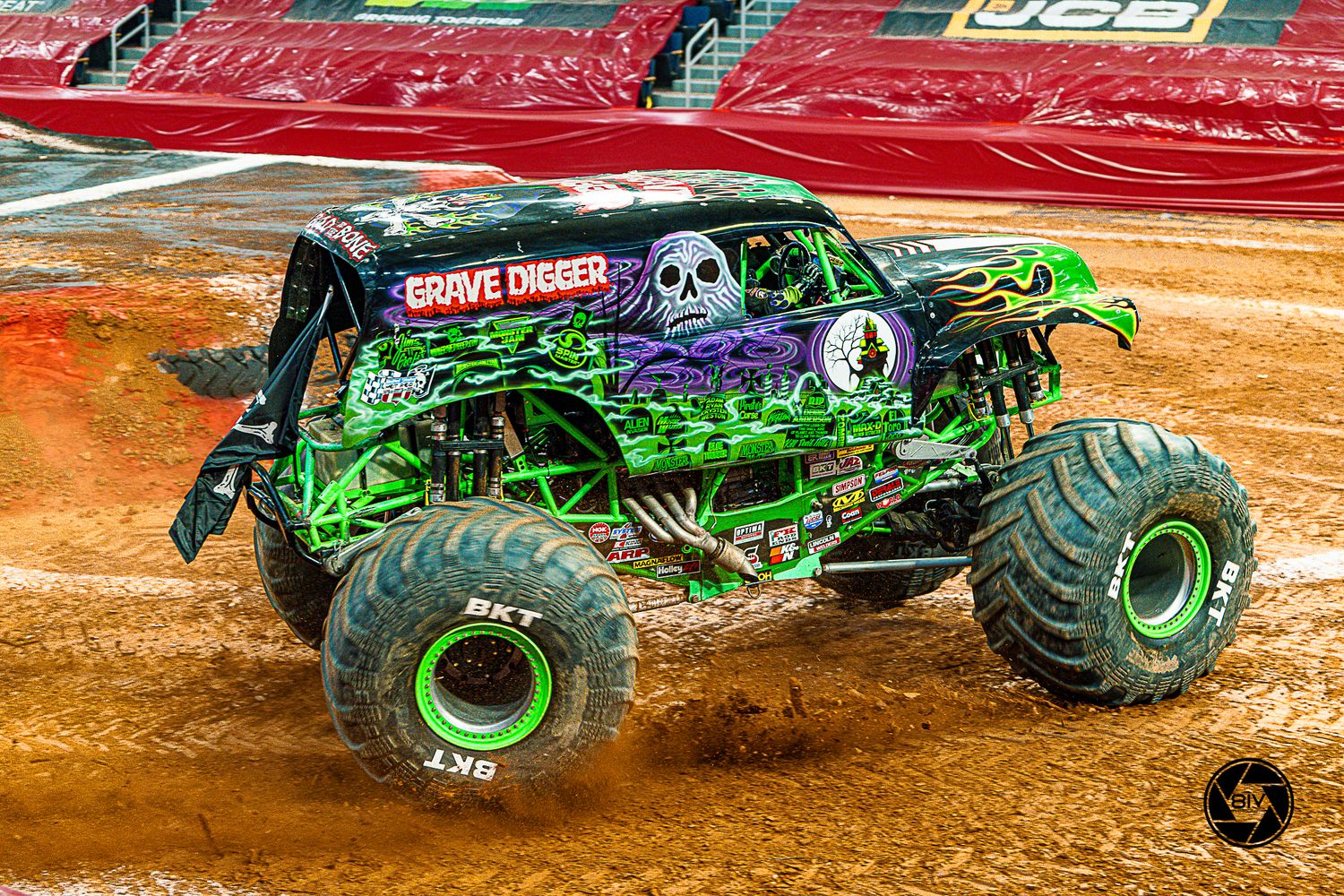 Grave Digger monster truck with green flames and skull design performs at an indoor arena on a dirt track.