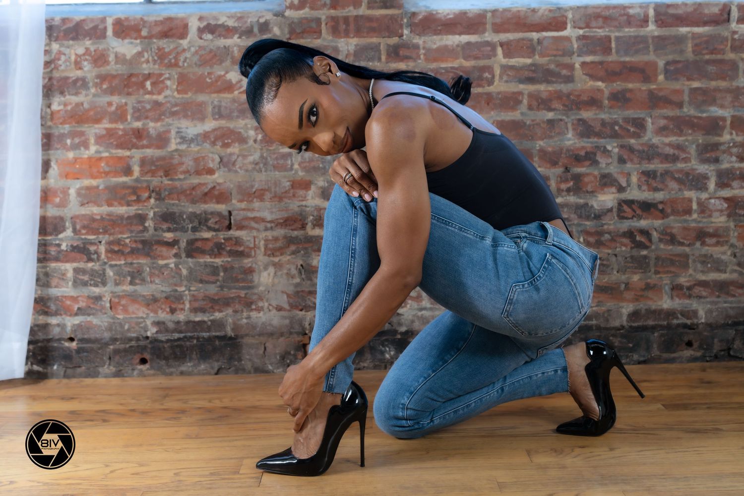 Woman in jeans and black stilettos posing against a brick wall in a professional photo shoot.