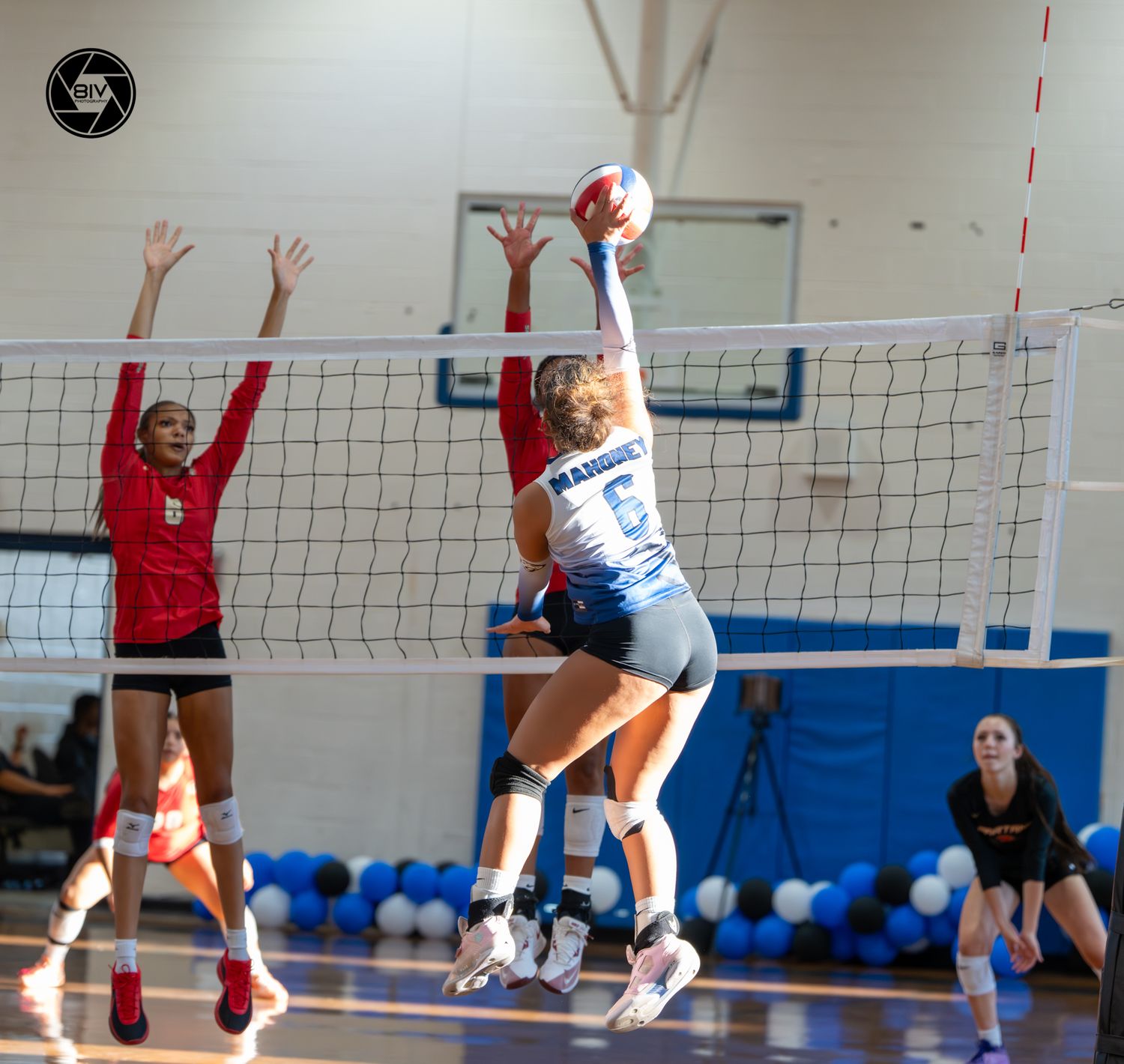 Female volleyball player in white uniform spikes ball over net against red-uniformed blocker during indoor match.