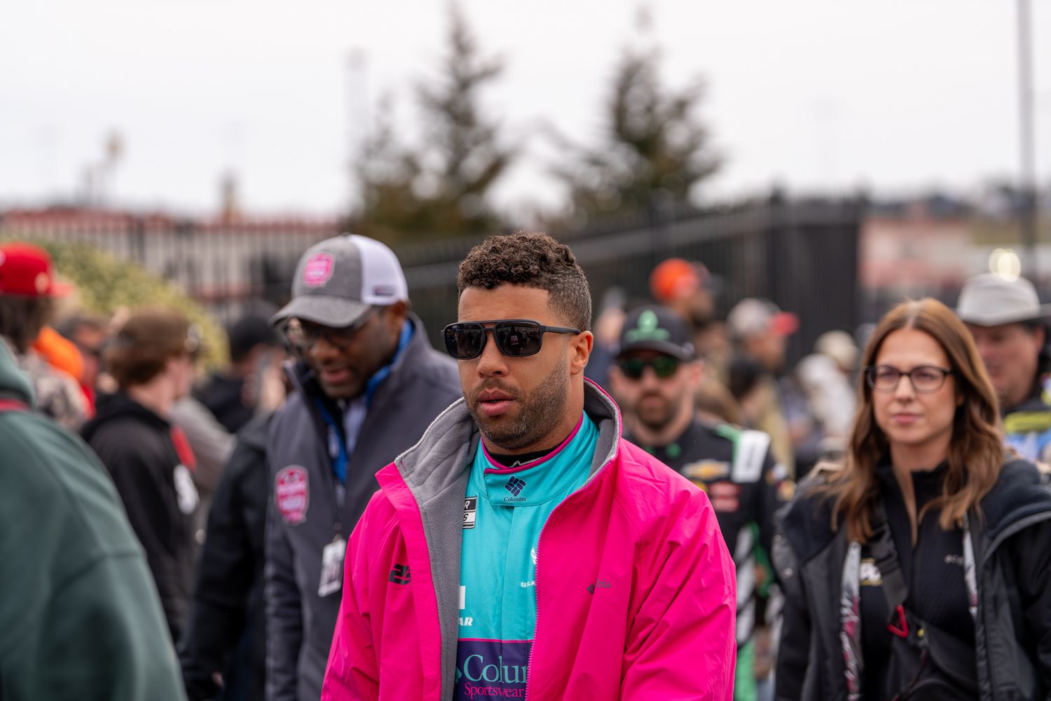 Bubba Wallace in pink jacket and sunglasses walks through outdoor crowd event on overcast day.