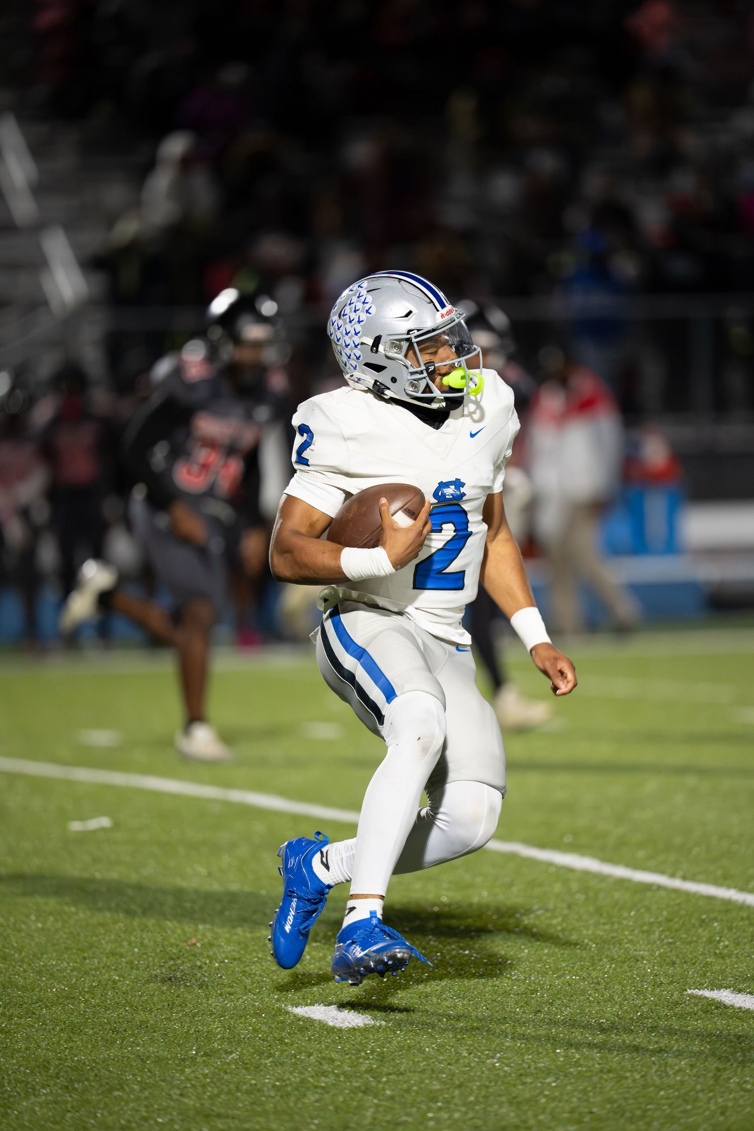 Football player #2 in white and blue uniform runs with the ball during a night game under stadium lights.