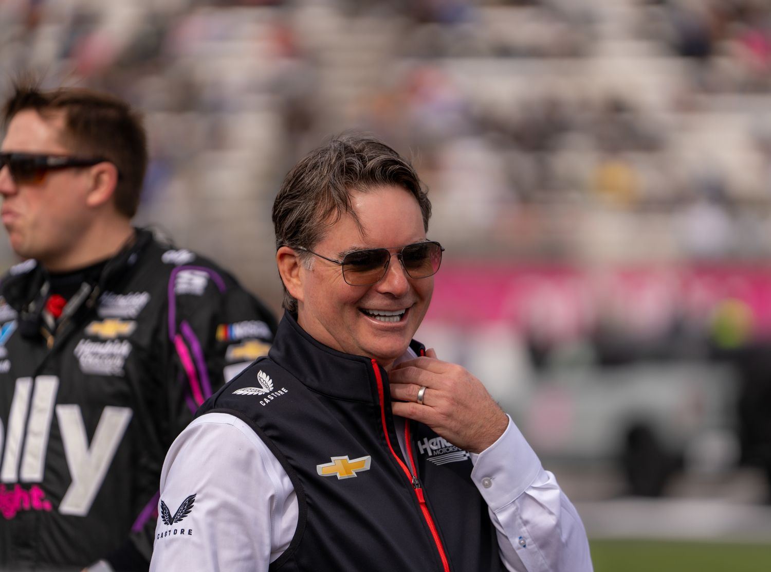 Jeff Gordon smiling at NASCAR race event wearing Chevrolet jacket and sunglasses with racing venue in background.