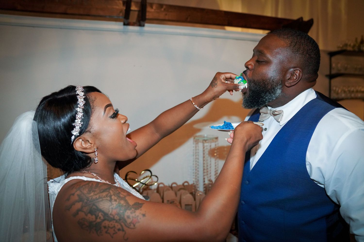 Bride feeding groom wedding cake during reception, bride wearing veil and groom in blue vest.