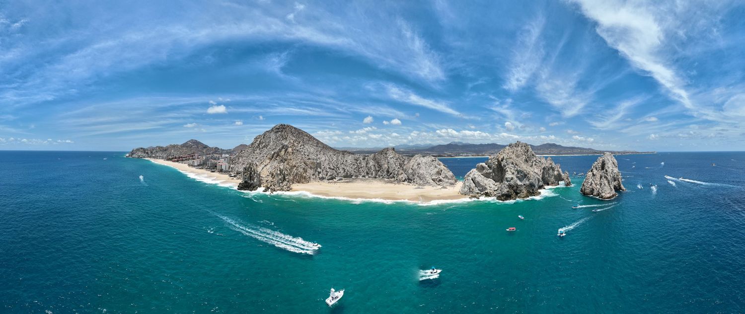 Aerial panoramic view of Cabo San Lucas coastline with turquoise waters, sandy beach, rocky cliffs, and boats under blue sky.