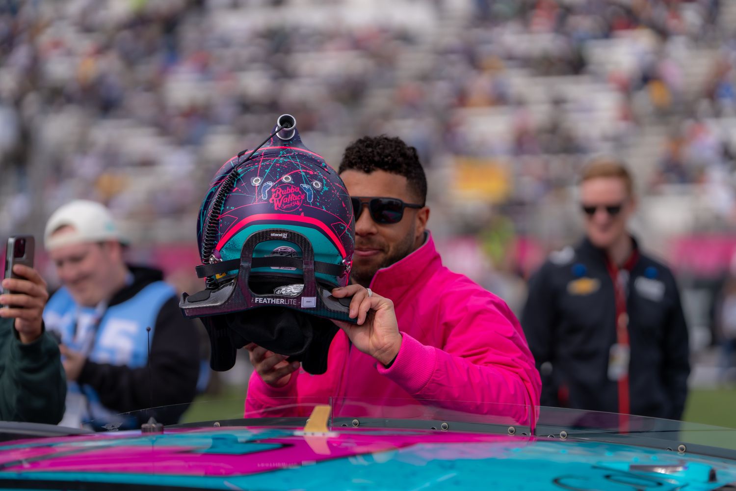 NASCAR driver, Bubba Wallace, in pink jacket holds decorated helmet at crowded racetrack event.