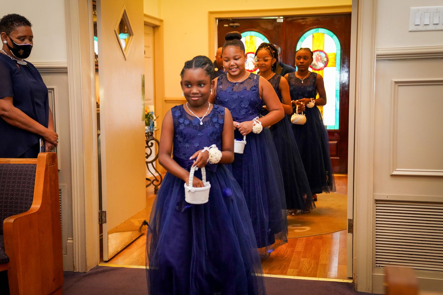 Young flower girls in navy blue tulle dresses walk down church aisle during wedding ceremony.
