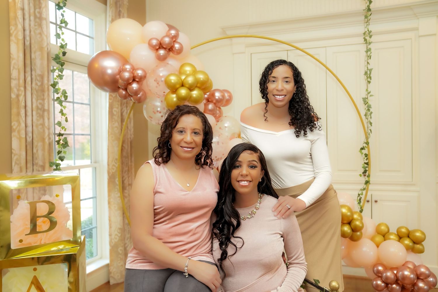 Three women pose together at a baby shower with pink, gold, and rose gold balloon decorations.