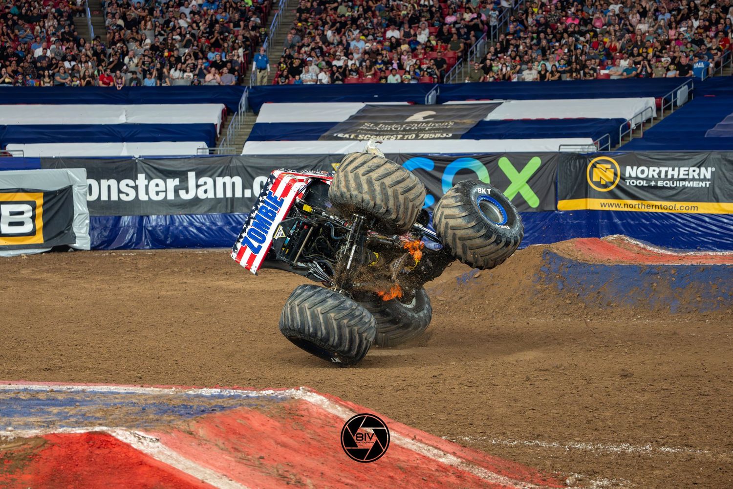 Zombie Monster Jam truck performing a dramatic wheelie stunt on a dirt track inside a packed stadium arena.