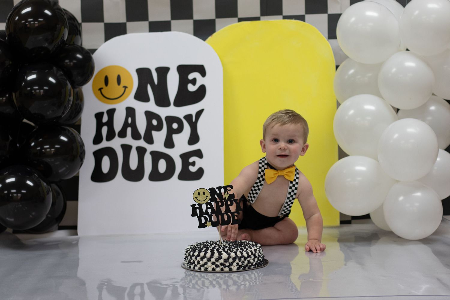 Baby boy in bow tie sits before One Happy Dude birthday backdrop with balloons and smash cake.