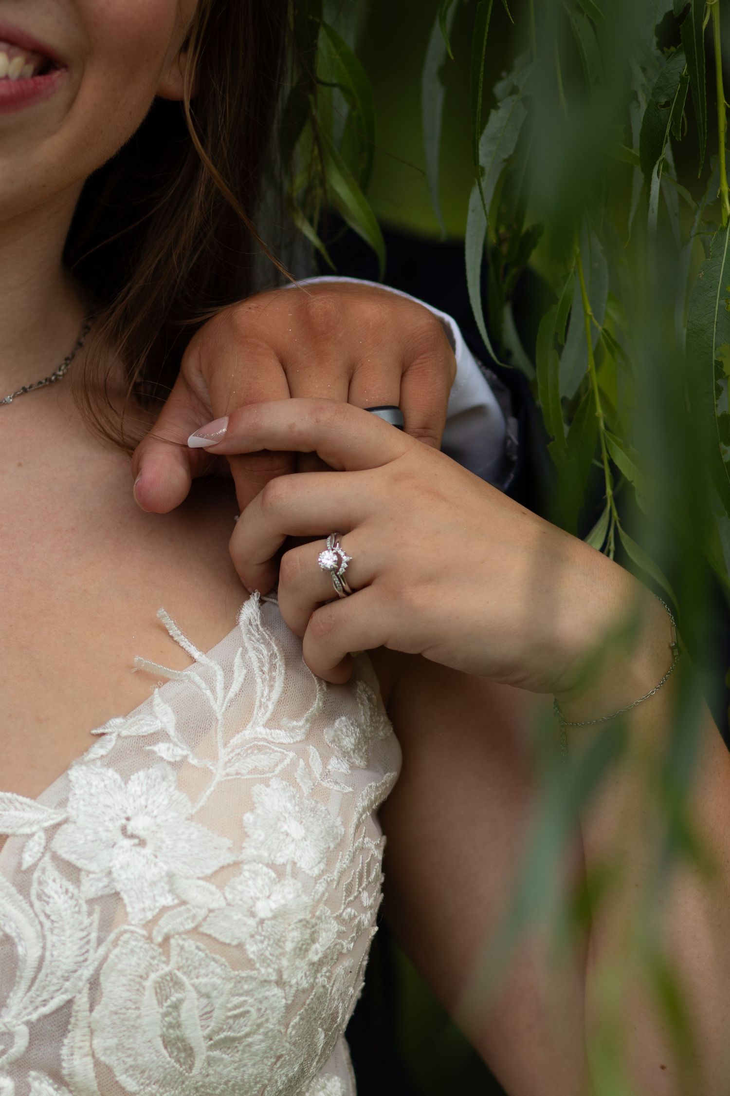 Close-up of a bride in lace wedding gown with diamond engagement ring among green leaves.