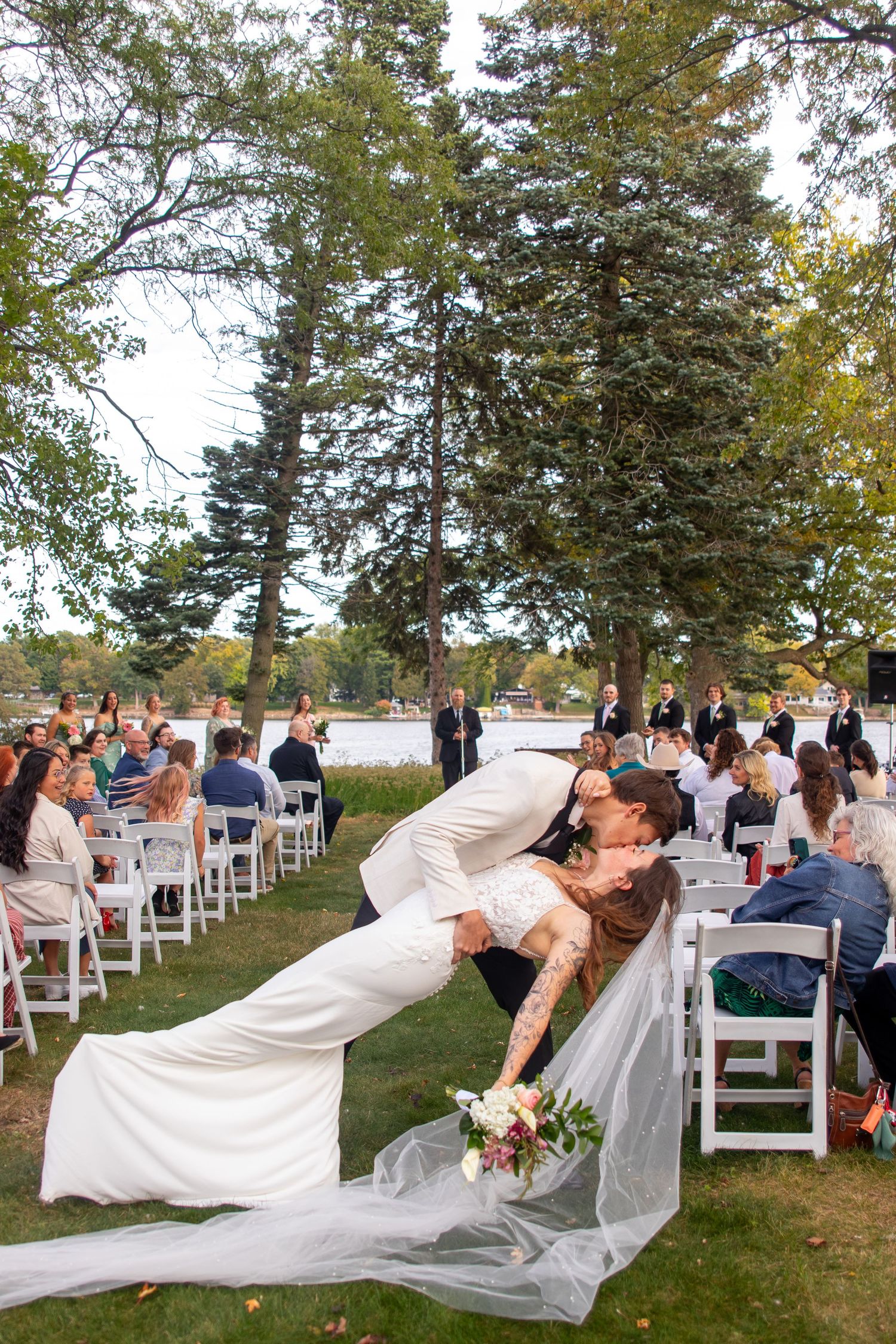 Bride and groom share a romantic dip kiss at outdoor wedding ceremony with guests seated on white chairs by a lakeside.