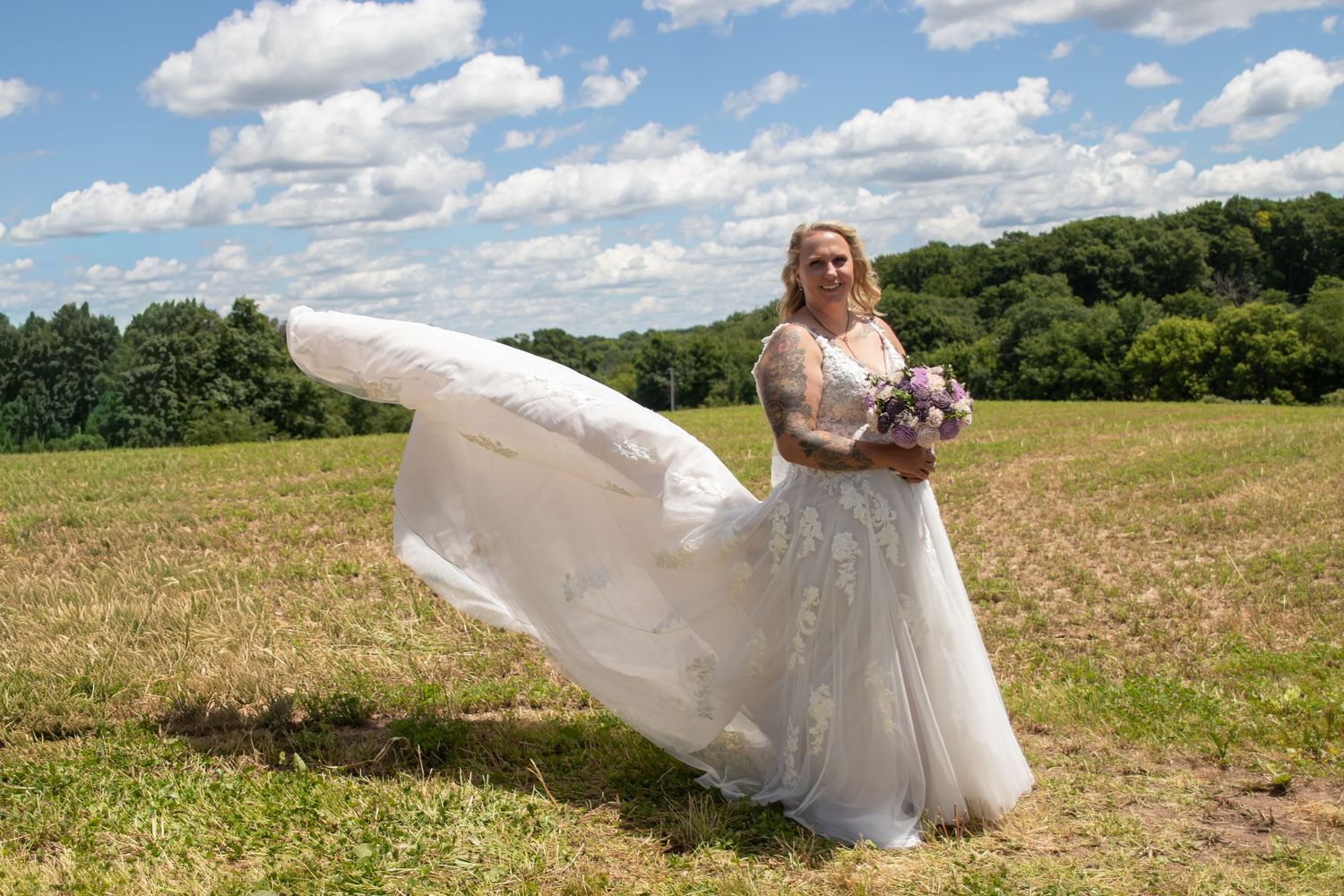 Bride in flowing white lace gown holding purple bouquet poses in open field with dramatic cloudy sky.