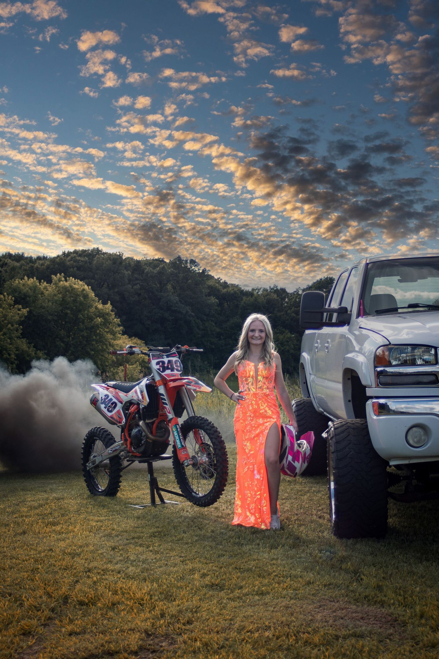 Senior girl in orange prom dress posing with dirt bike and lifted truck at golden hour sunset.