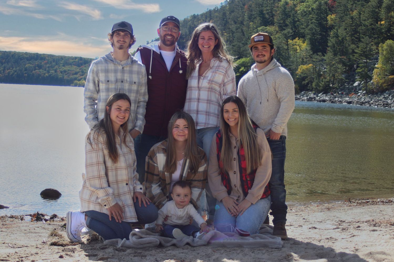 Family of eight poses together on sandy beach shore with scenic lake and forested hills in background.