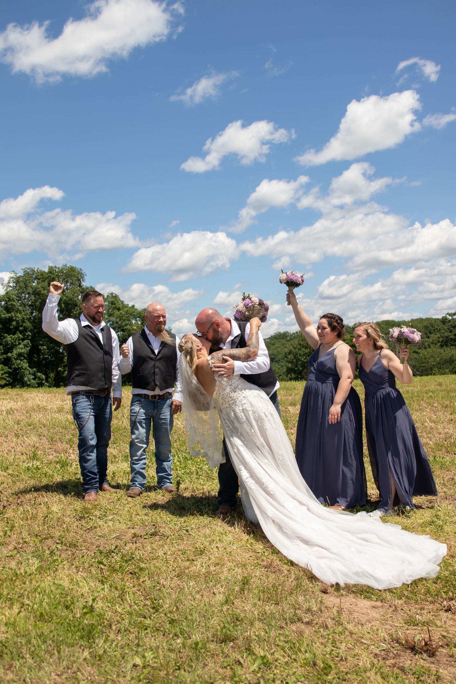 Bride and groom share a romantic kiss surrounded by groomsmen and bridesmaids in blue dresses on a sunny outdoor wedding day.