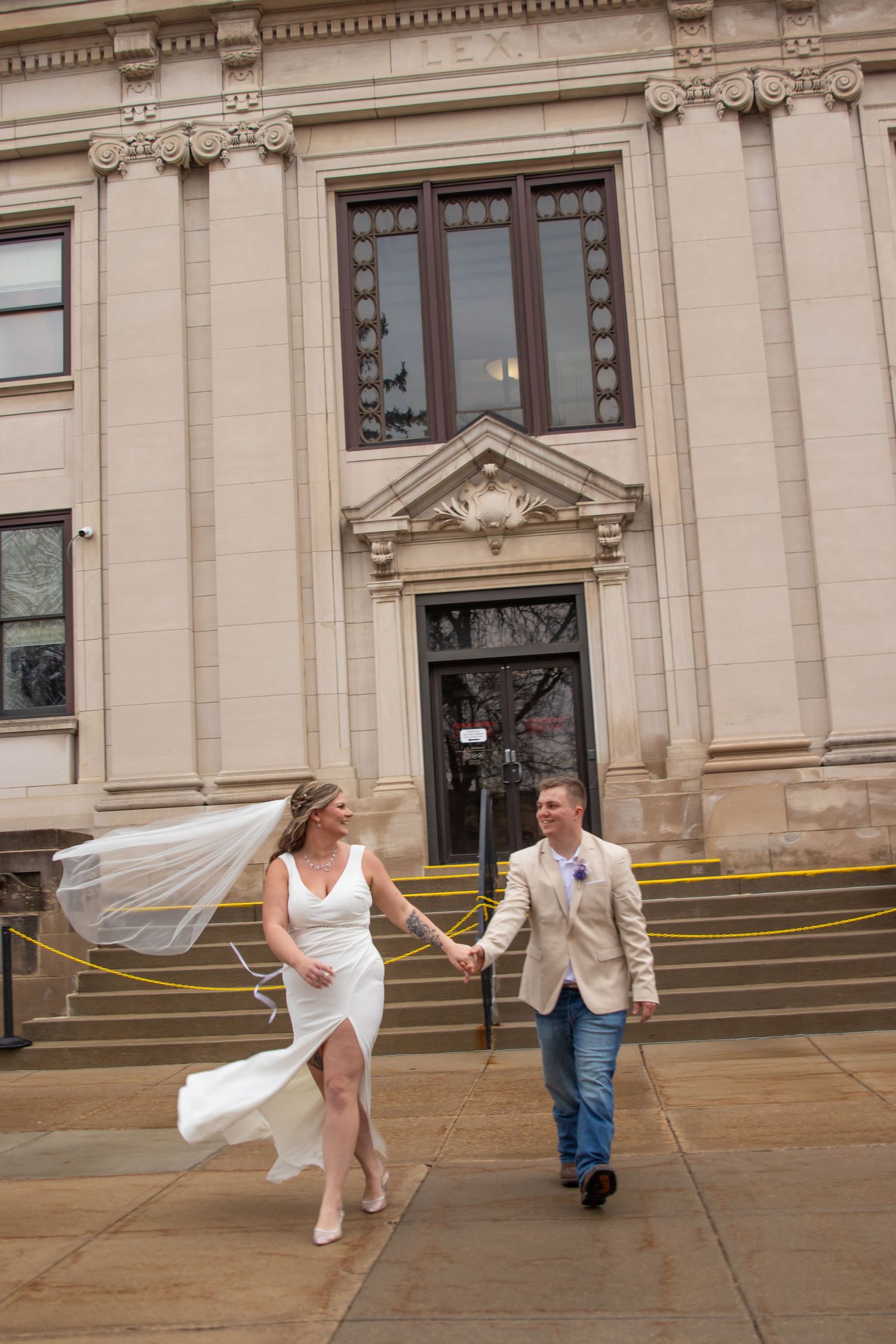 Bride and groom holding hands walking outside a classic stone building, veil blowing in the wind on their wedding day.