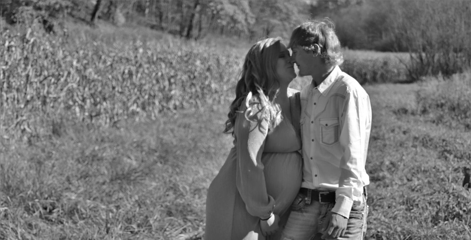 Black and white image of expectant couple kissing gently in a sunlit rural countryside field.