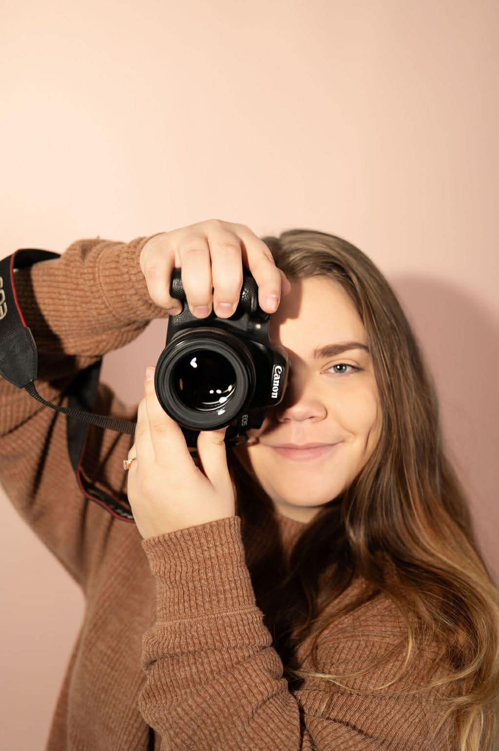 Woman in brown sweater holding Canon DSLR camera up to her face against pink background.