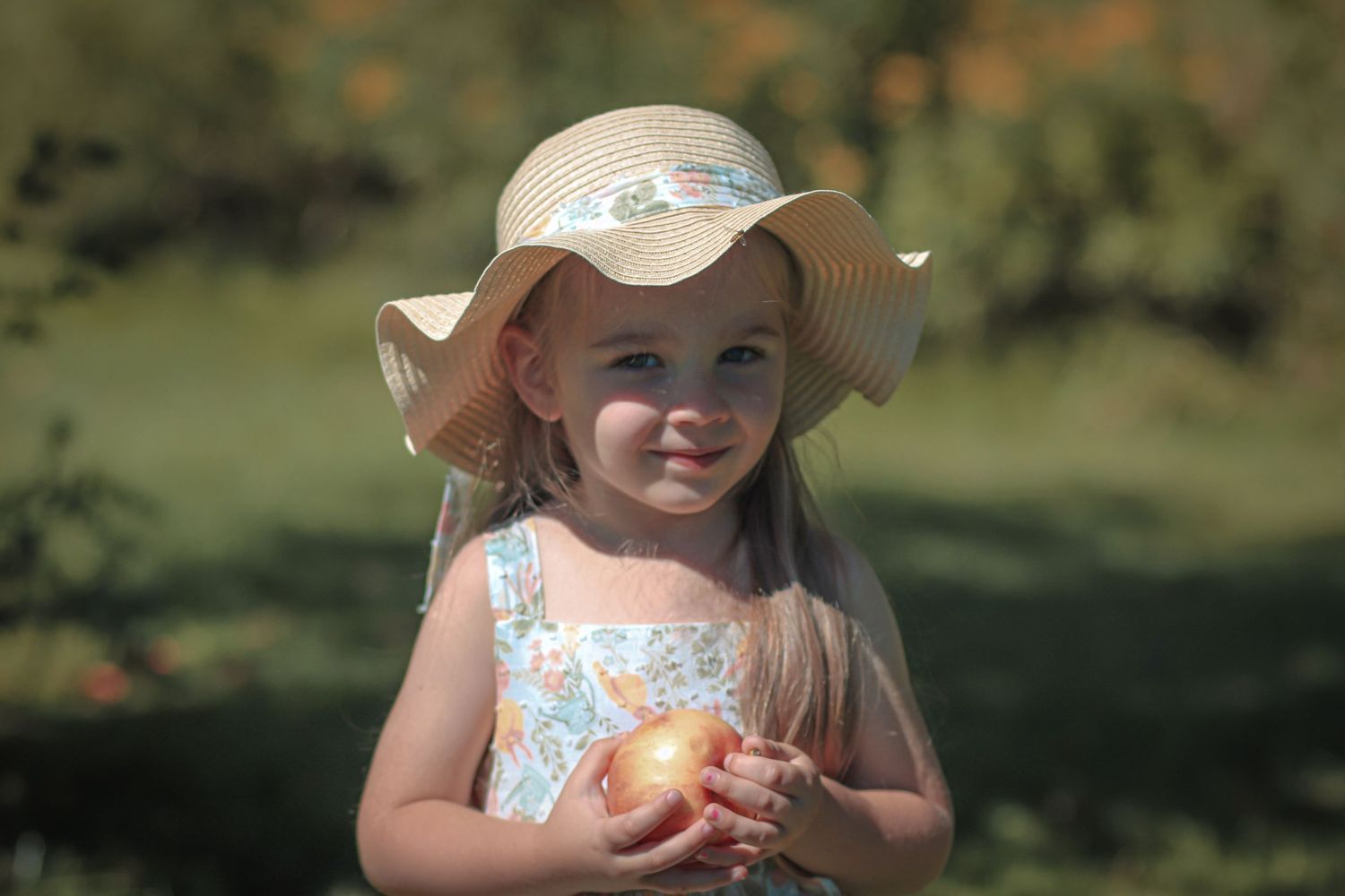 A young girl in a floral dress and wide-brim sun hat smiles while holding a fresh apple outdoors.
